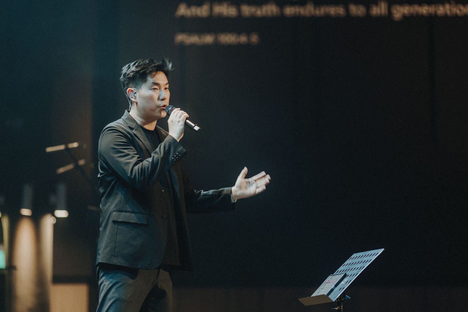 A performer sings passionately at a worship service with a Bible verse displayed above on a dark background.