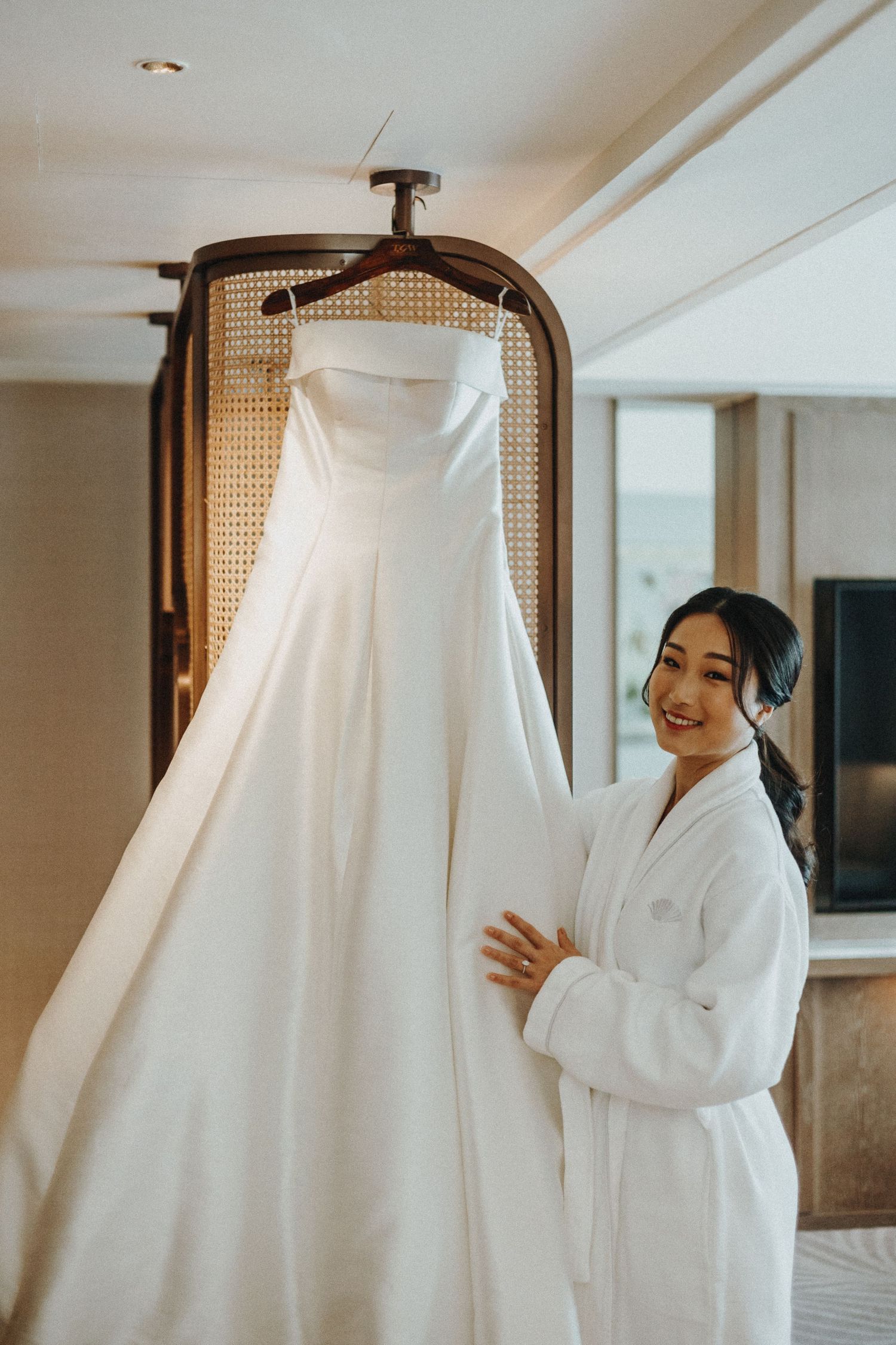 A beautiful white wedding dress hangs on display while someone in a white robe admires it in a hotel room.