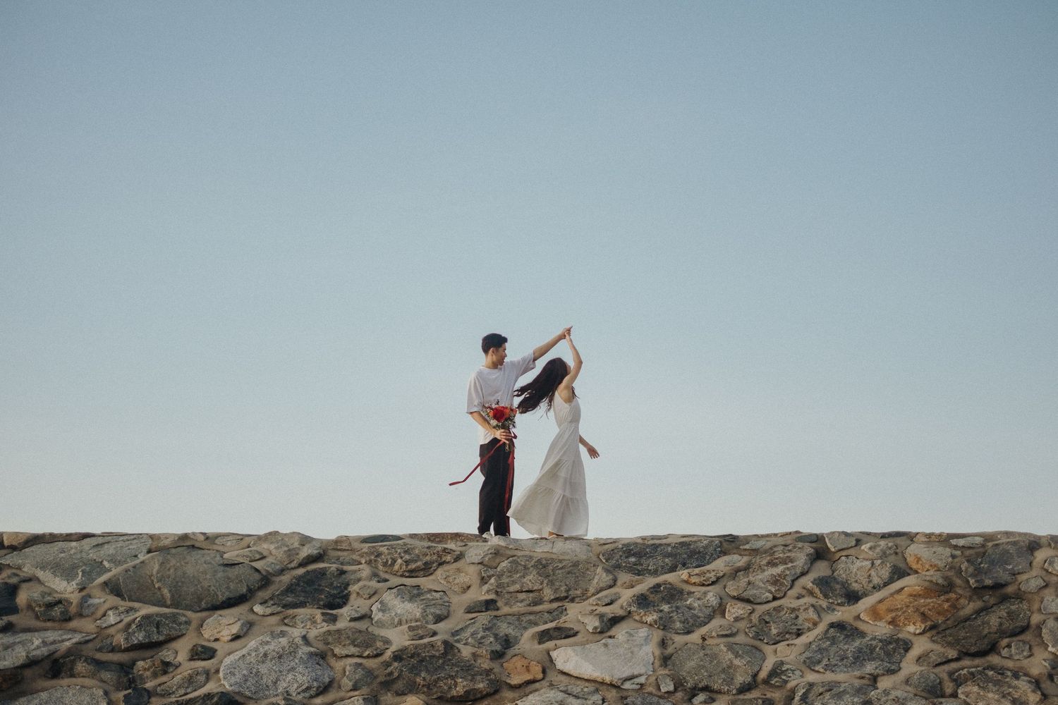 Two people stand together on a rocky seaside cliff against a pale blue sky.