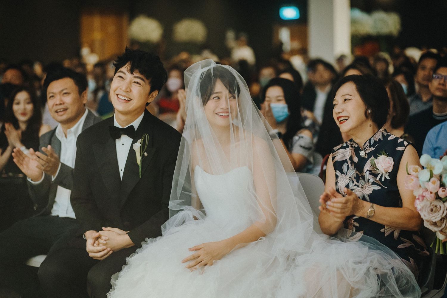 Wedding guests smile and applaud during an indoor ceremony as the bride in white veil sits among the audience.