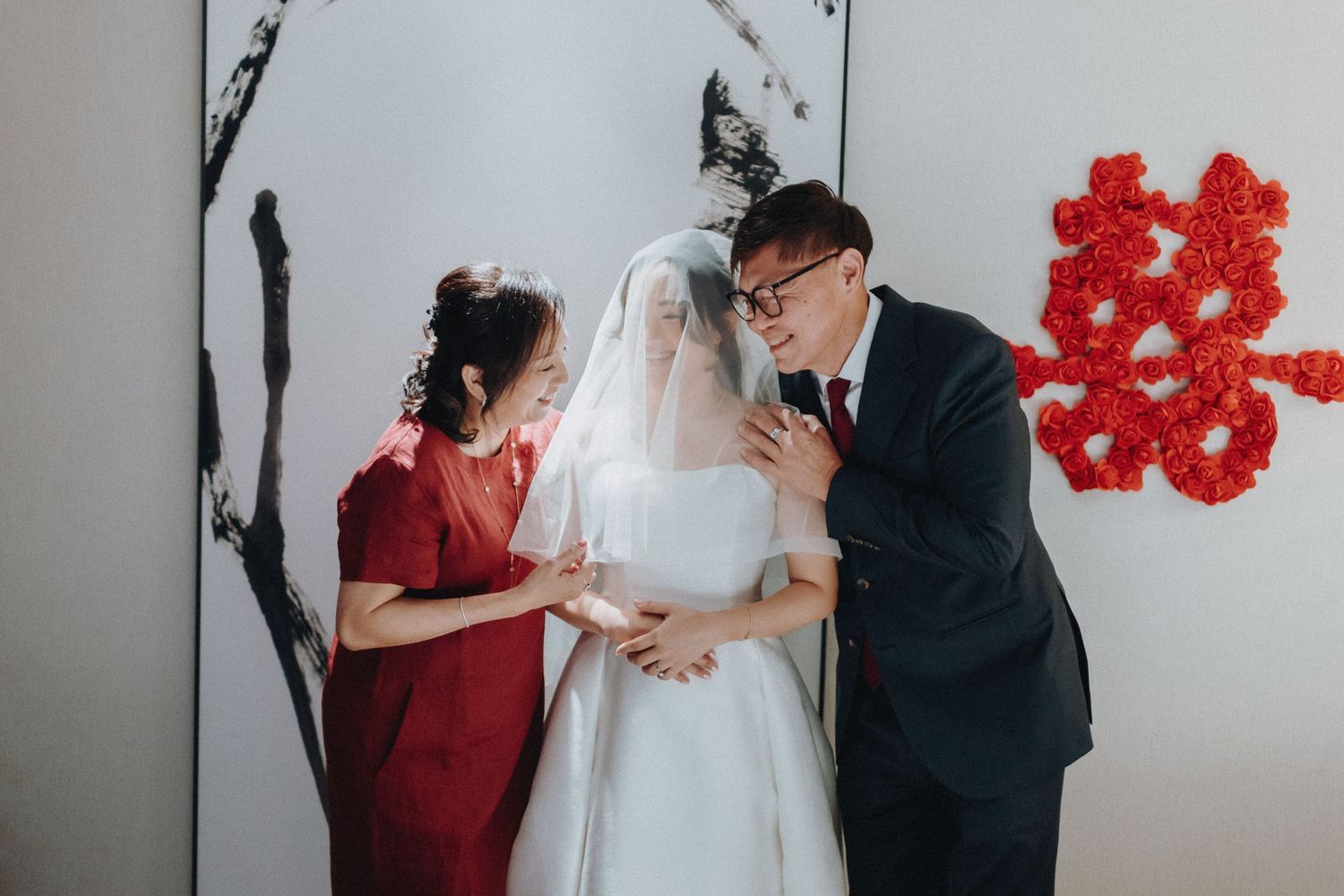 A wedding couple and guest share an intimate moment with red floral decorations in the background.