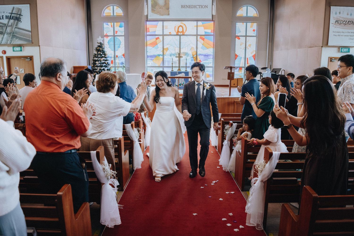 Newlyweds walk down the red carpet aisle as guests applaud in a decorated church.