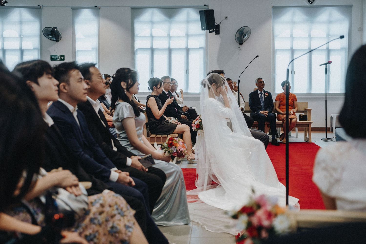 Wedding ceremony in a church with bride in white gown and veil seated among guests in formal attire.