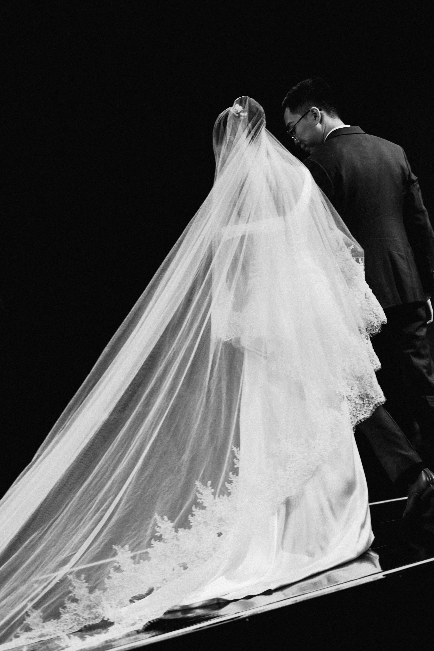 Black and white photo of bride and groom walking, featuring elegant lace cathedral veil trailing behind wedding dress.