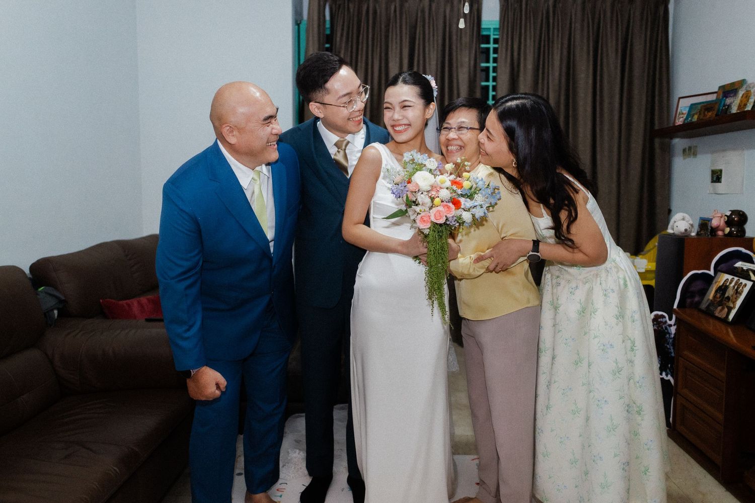 Happy wedding couple with bridal bouquet shares joyful moment with family members in casual home setting.
