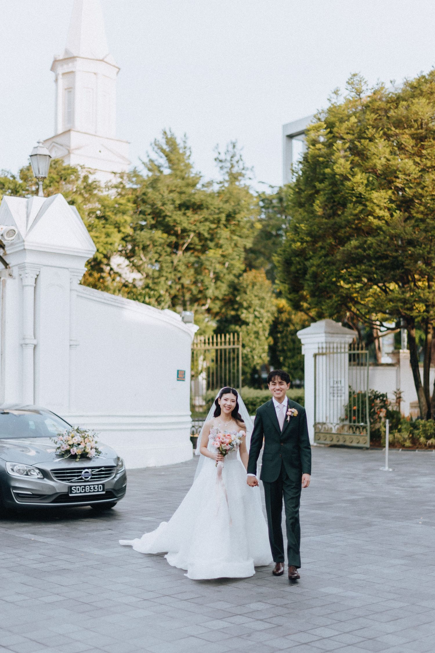 Bride and groom walking hand in hand outside white church building surrounded by green trees.