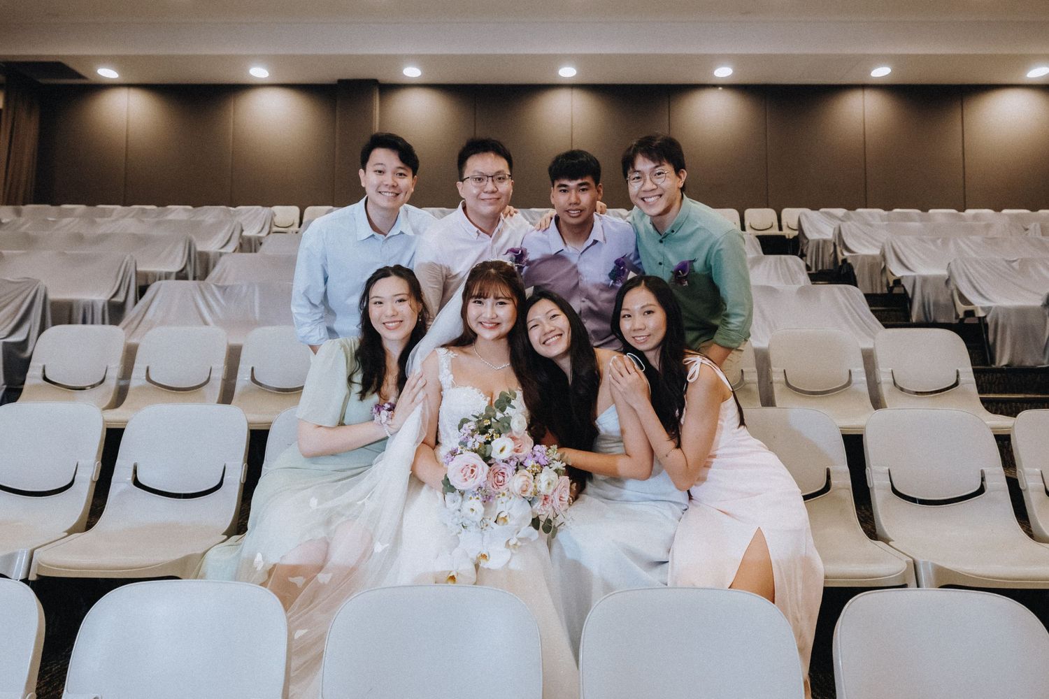 Wedding party members pose together in white formal attire on auditorium seats.