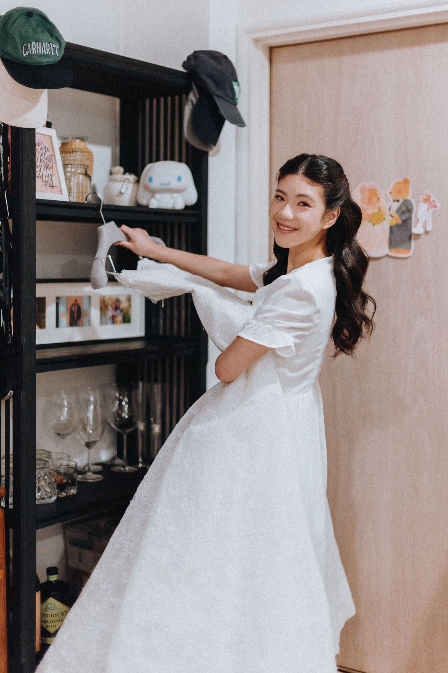 Young Asian woman in white dress smiling while reaching for items on a dark bookshelf in a cozy room.