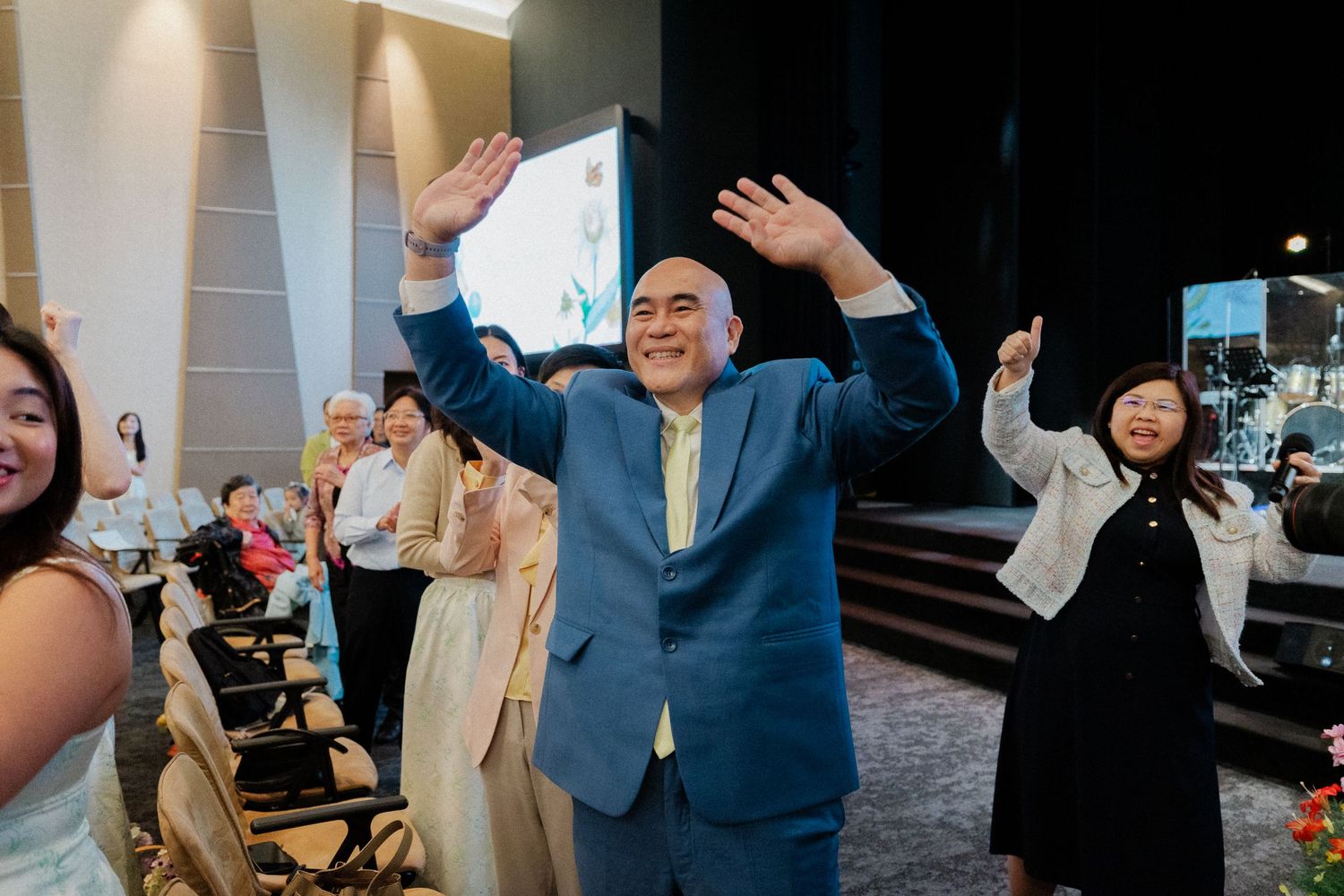Man in blue suit raises hands joyfully at wedding celebration with guests cheering around him.