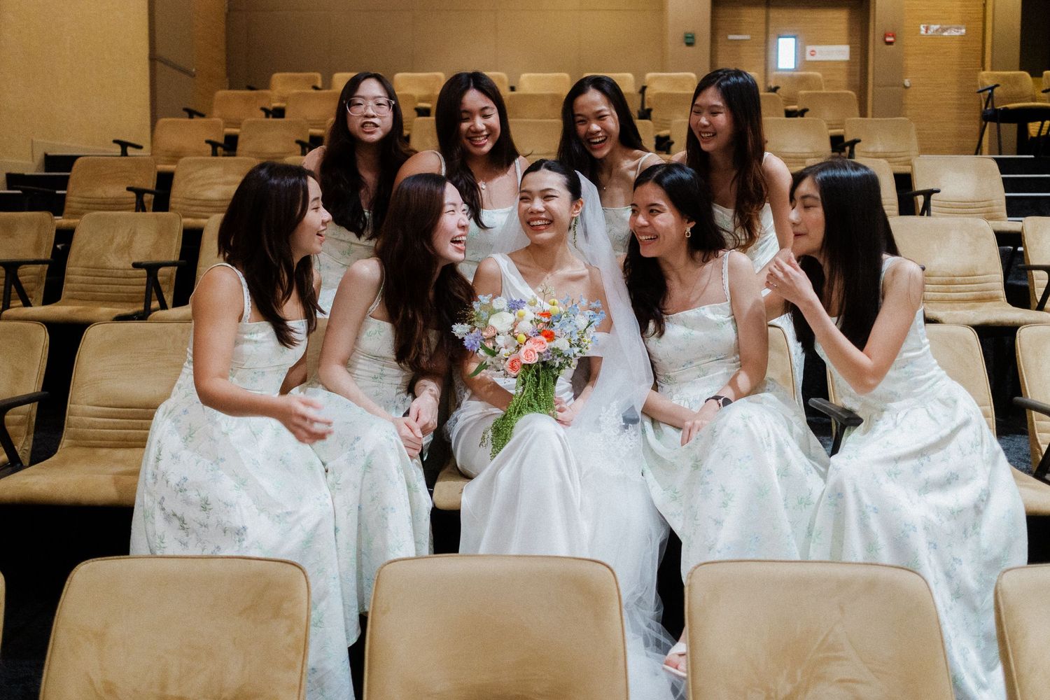 Bride surrounded by bridesmaids in white dresses, holding colorful bouquet, seated in auditorium chairs.