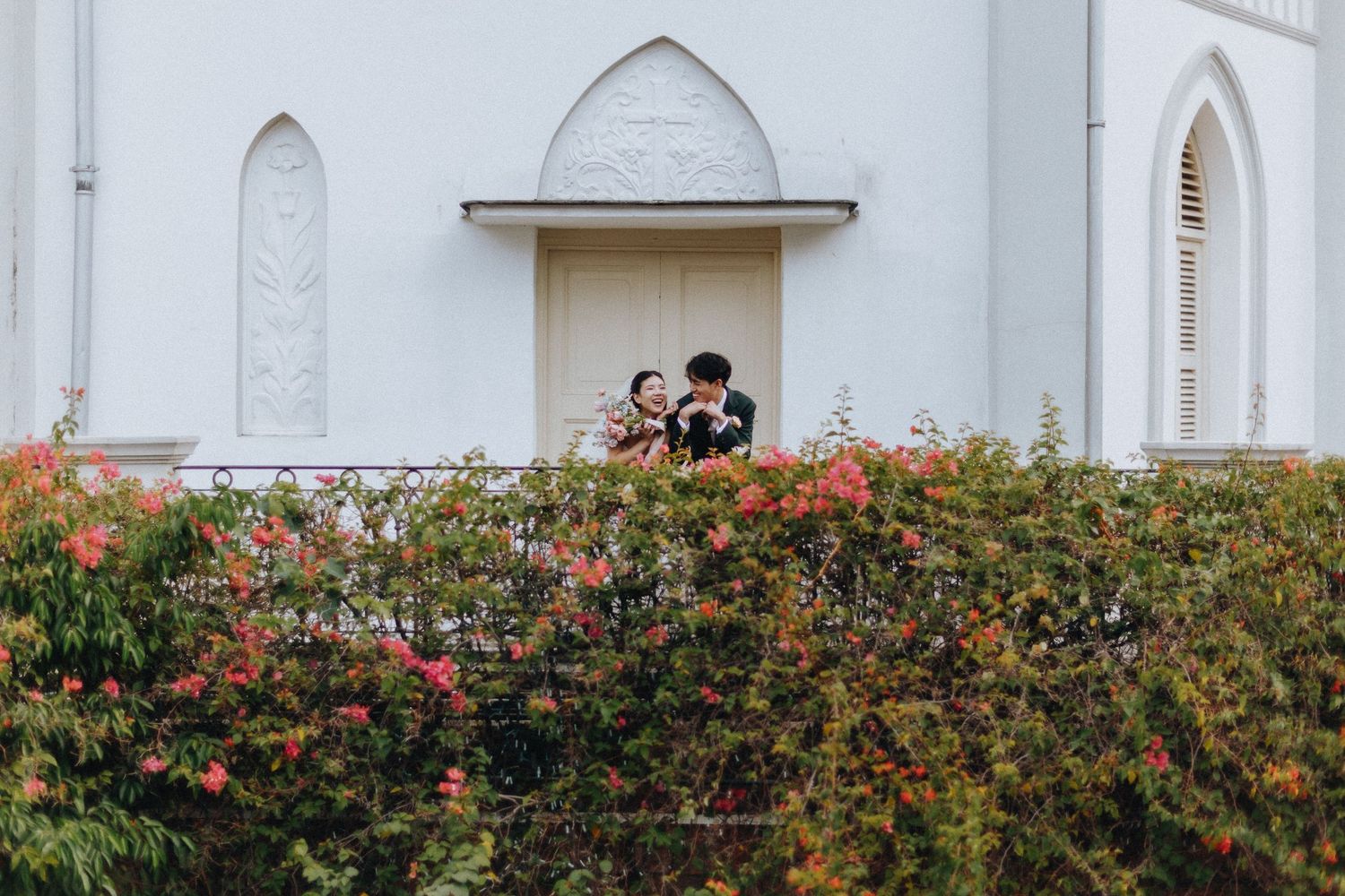 Couple embracing on a white church balcony surrounded by lush green and red flowering hedges.