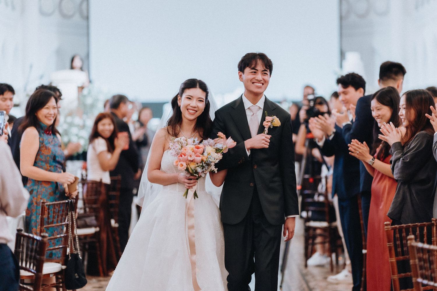 Happy newlywed Asian couple walks down the aisle at an elegant wedding ceremony with guests applauding.