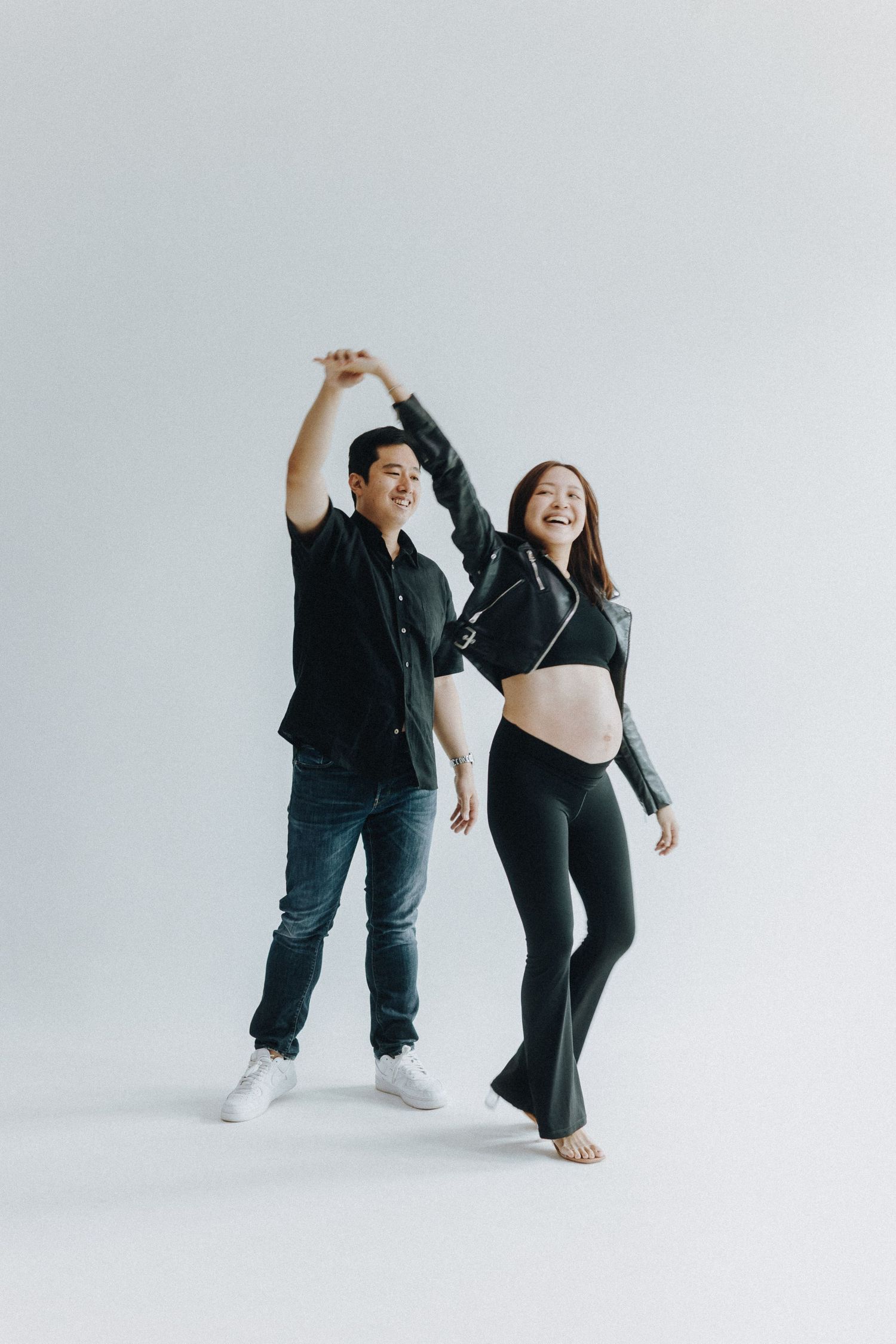 Dancing couple in casual wear, with pregnant woman in black outfit spinning against white background.