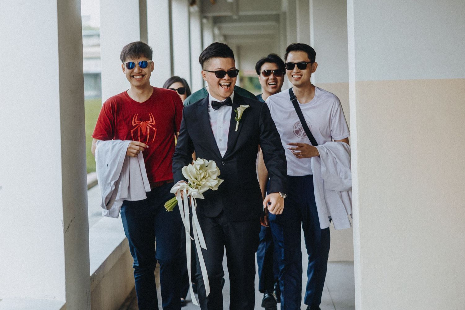 Group of groomsmen wearing sunglasses and formal attire pose together in a bright hallway.