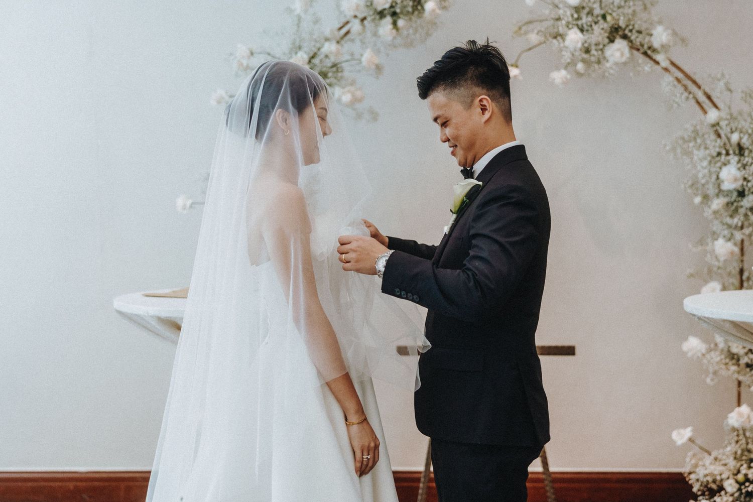 A couple in formal wedding attire sharing a tender moment under floral decorations at their ceremony.