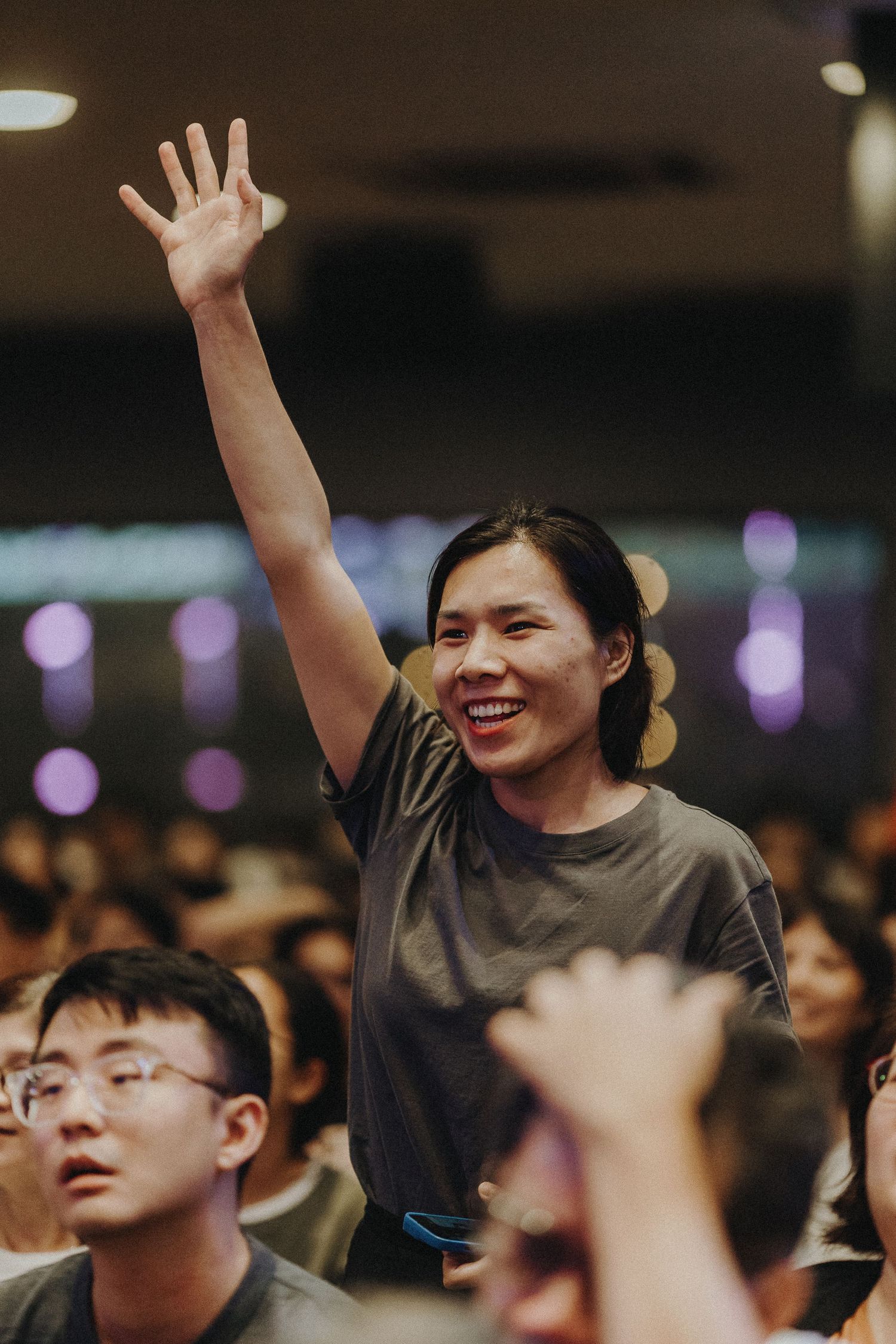 Audience members raise their hands in excitement at a concert venue with purple lighting overhead.