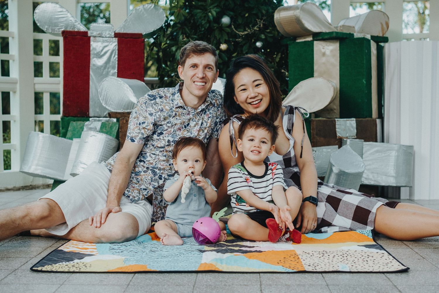 A family sits together on a picnic blanket outdoors with white balloons and red and green decorations in the background.