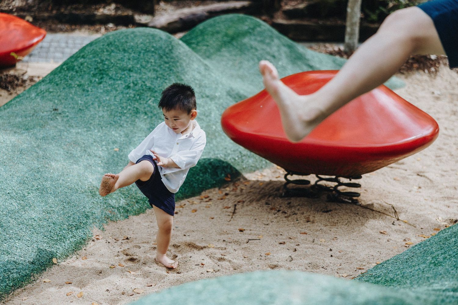 Children play at a playground with a red spinning disc and green undulating ground surface.