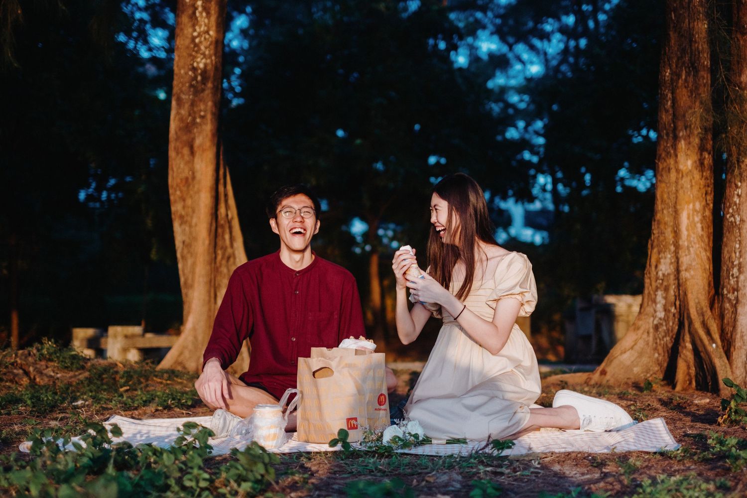 Two people sharing a picnic on a blanket under tall trees at sunset.