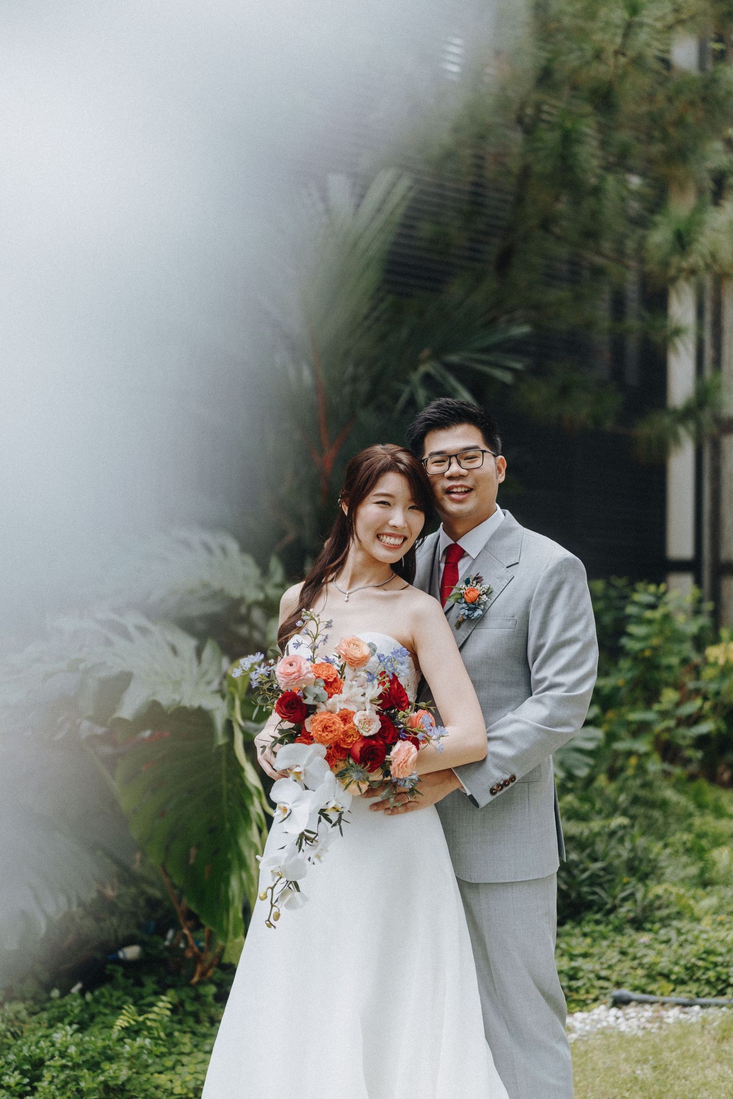 A couple poses together in their wedding attire surrounded by misty garden greenery.