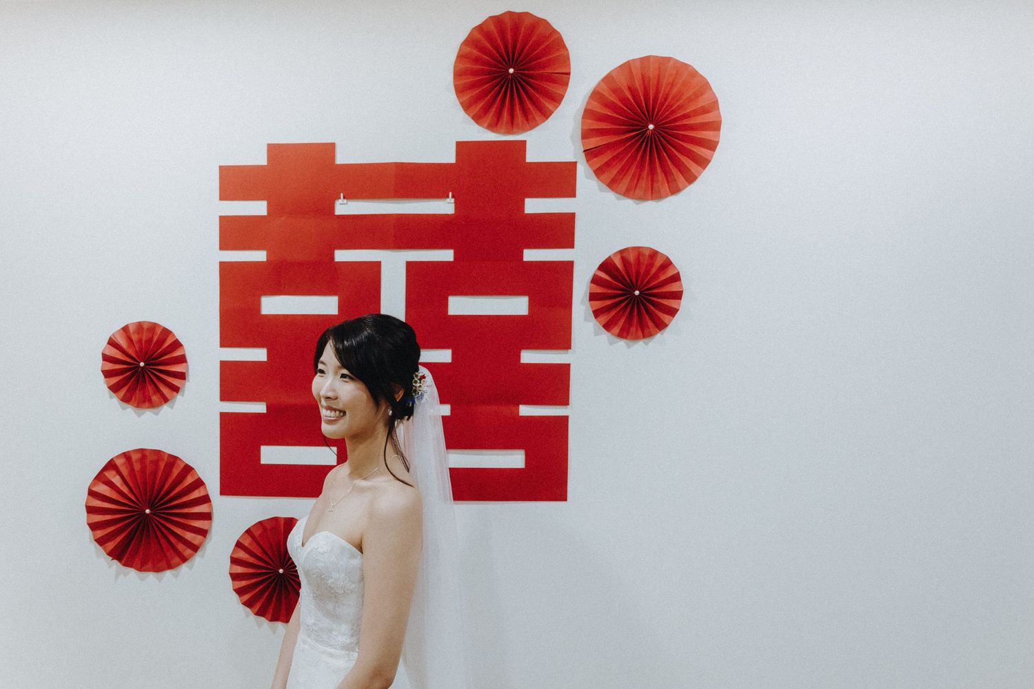 A bride in white dress stands against white wall with red double happiness symbol and paper fans for Chinese wedding decor.