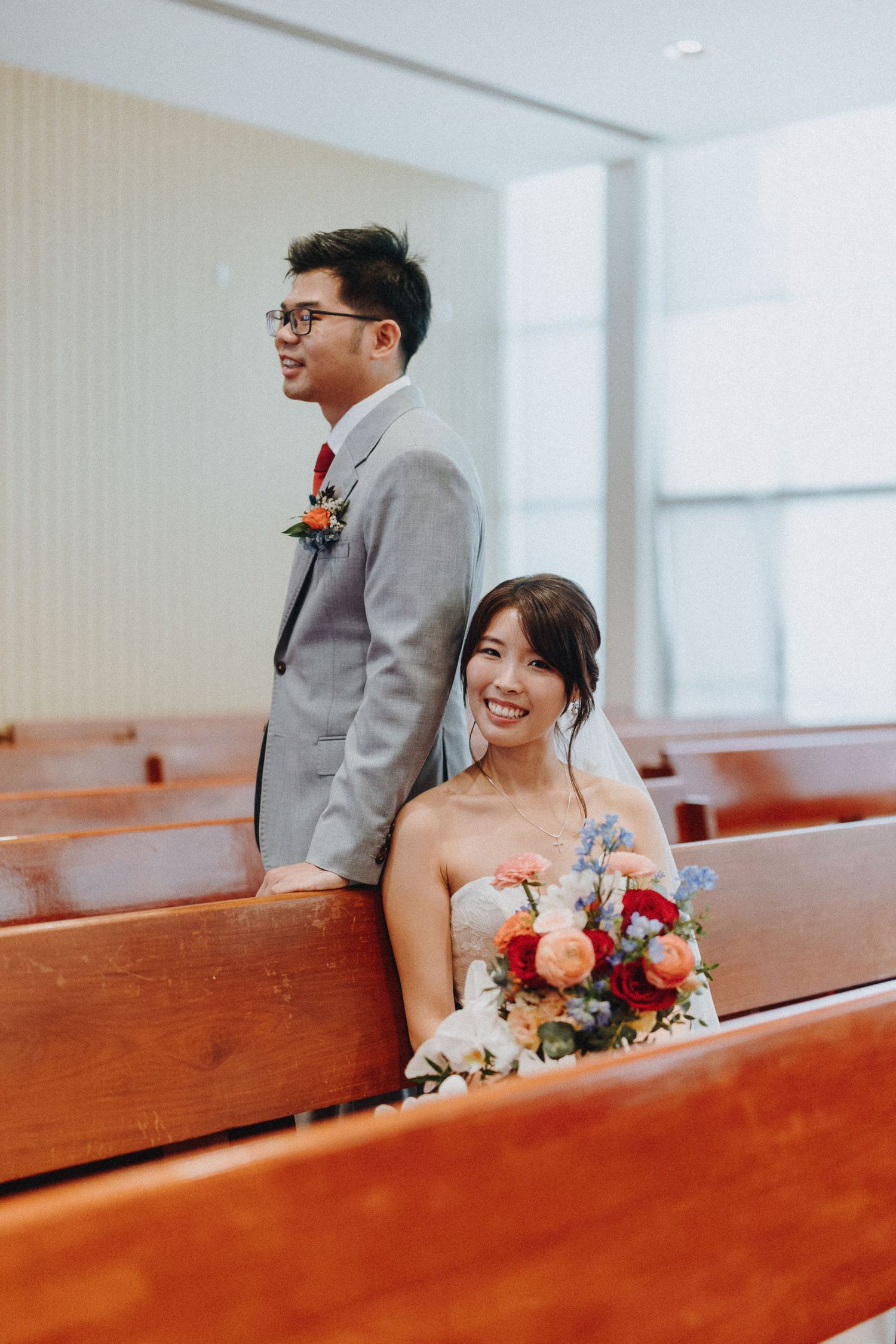 Newlyweds share a candid moment on wooden church pews, with the bride holding a vibrant bouquet.