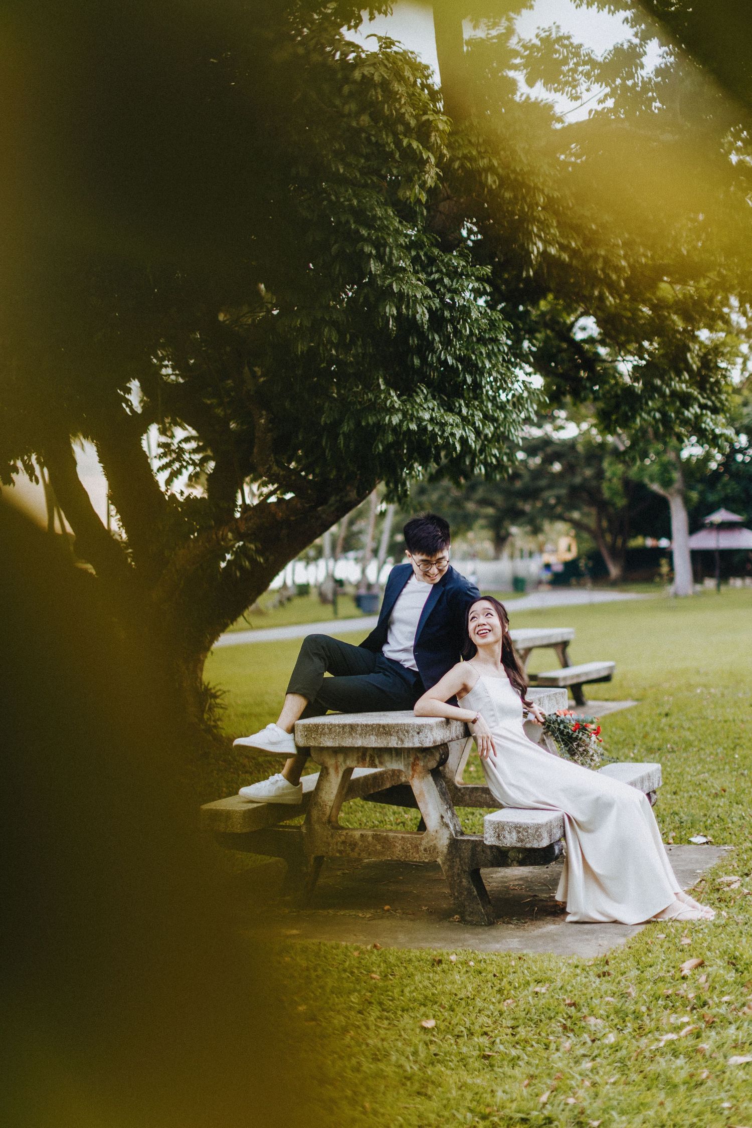 Romantic couple relaxing on a wooden bench in a sunlit park setting wearing formal attire.