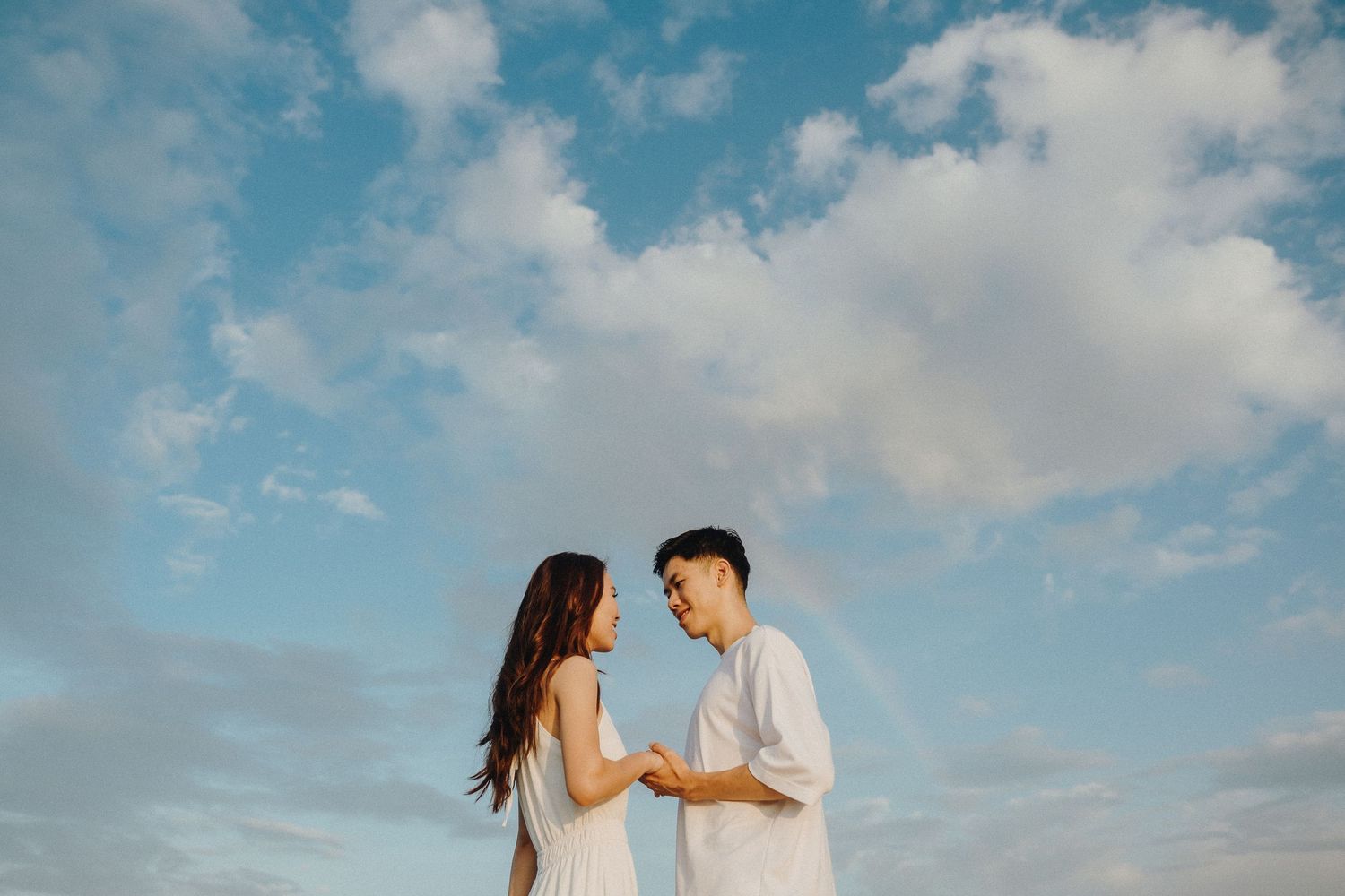 A couple shares tender moments against a dreamy blue sky with clouds during a romantic outdoor photoshoot.
