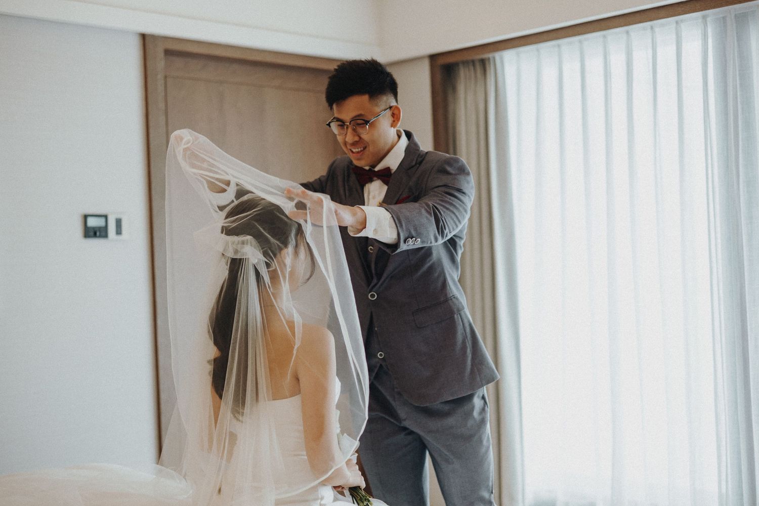 First look moment captured in a bright hotel room with flowing white curtains.