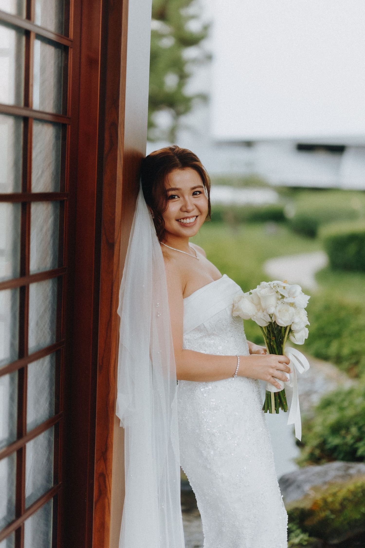 Bride in white lace dress poses with bouquet by wooden window frame.