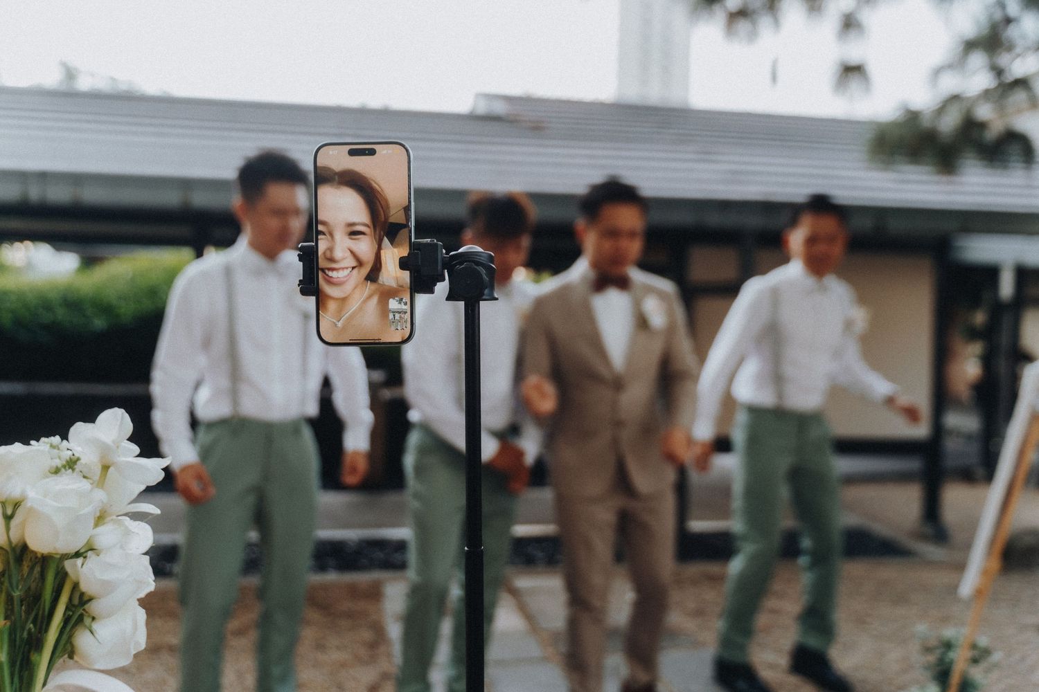 A smartphone mounted on a tripod captures people standing outside, with white flowers in the foreground.