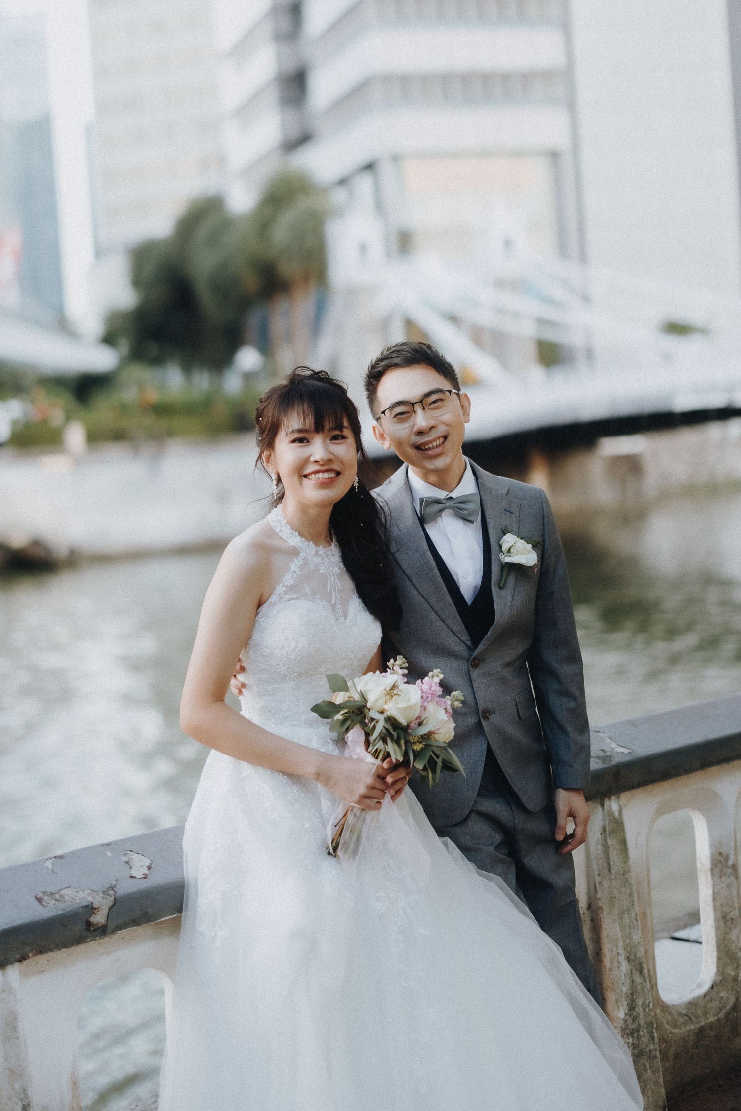 A romantic wedding couple poses along a waterfront with high-rise buildings in the background.
