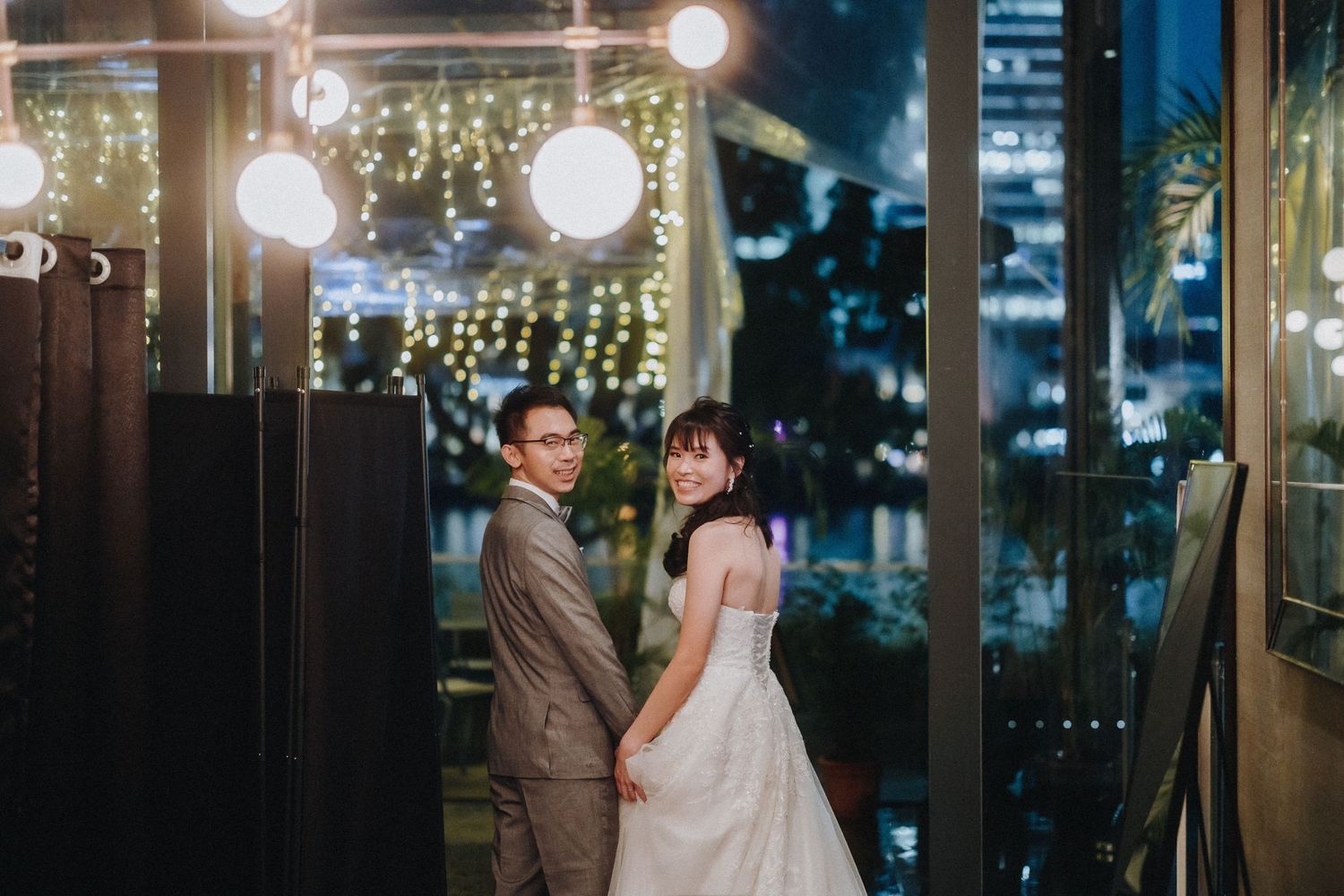 A couple in formal wedding attire hold hands beneath glowing string lights and lanterns during a romantic evening celebration.