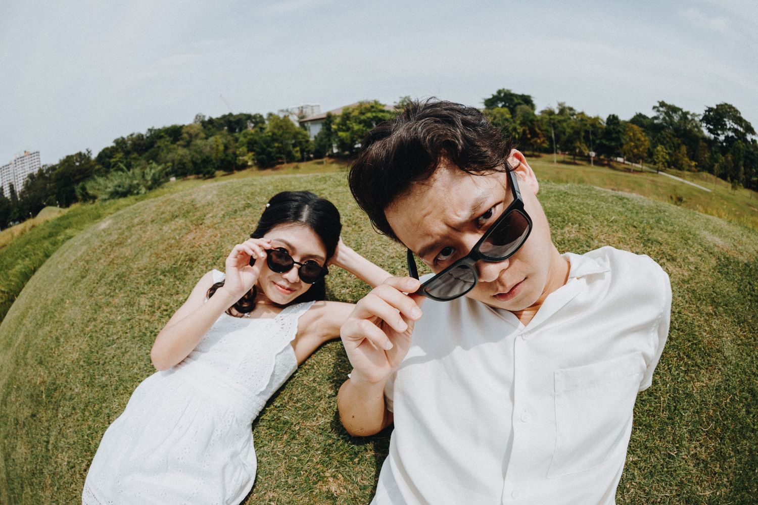 A couple in white clothing poses playfully with sunglasses in a fisheye lens photo on a grassy hillside.
