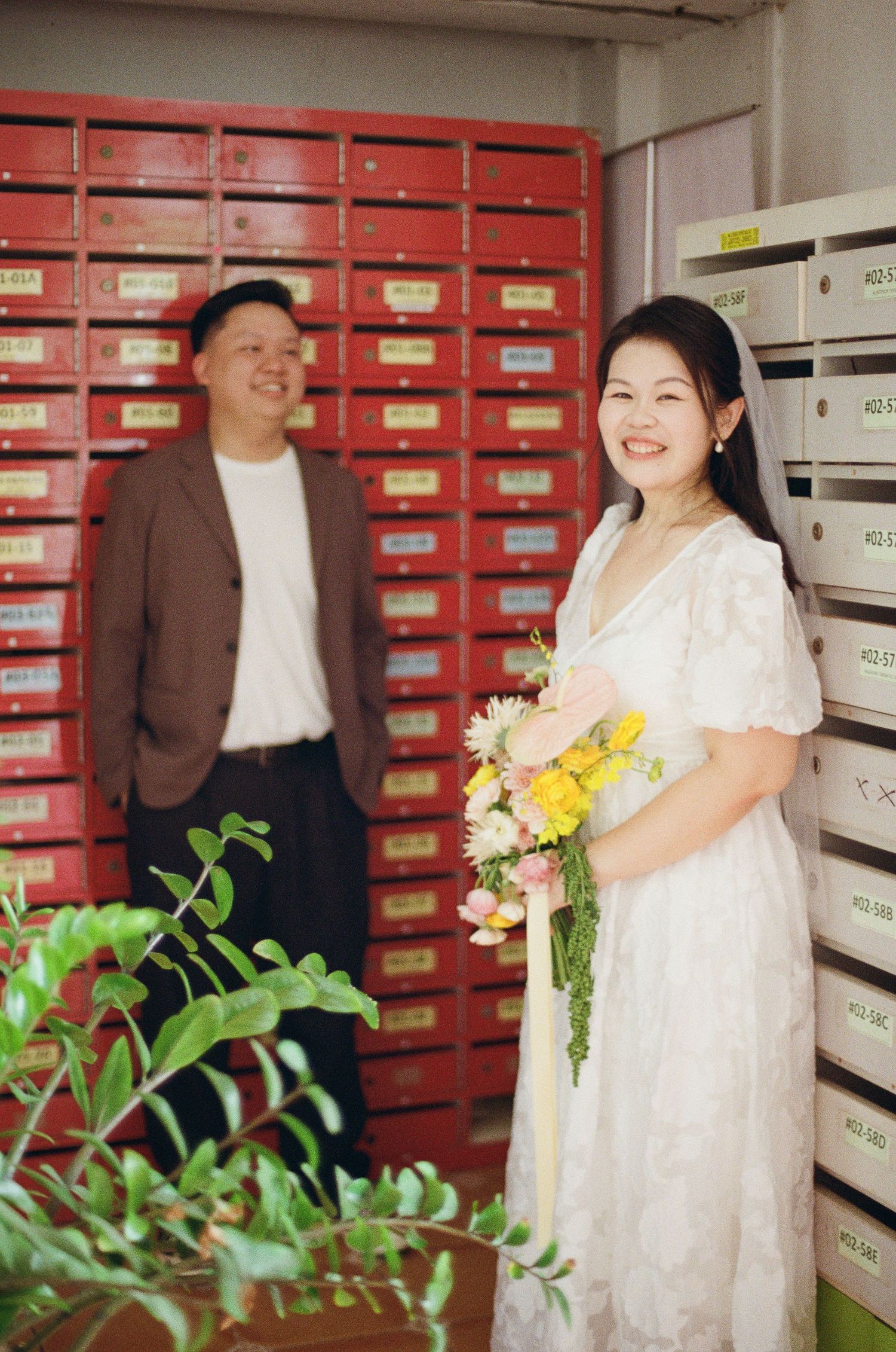 A couple poses playfully together in a mail room with red mailboxes and flowers.