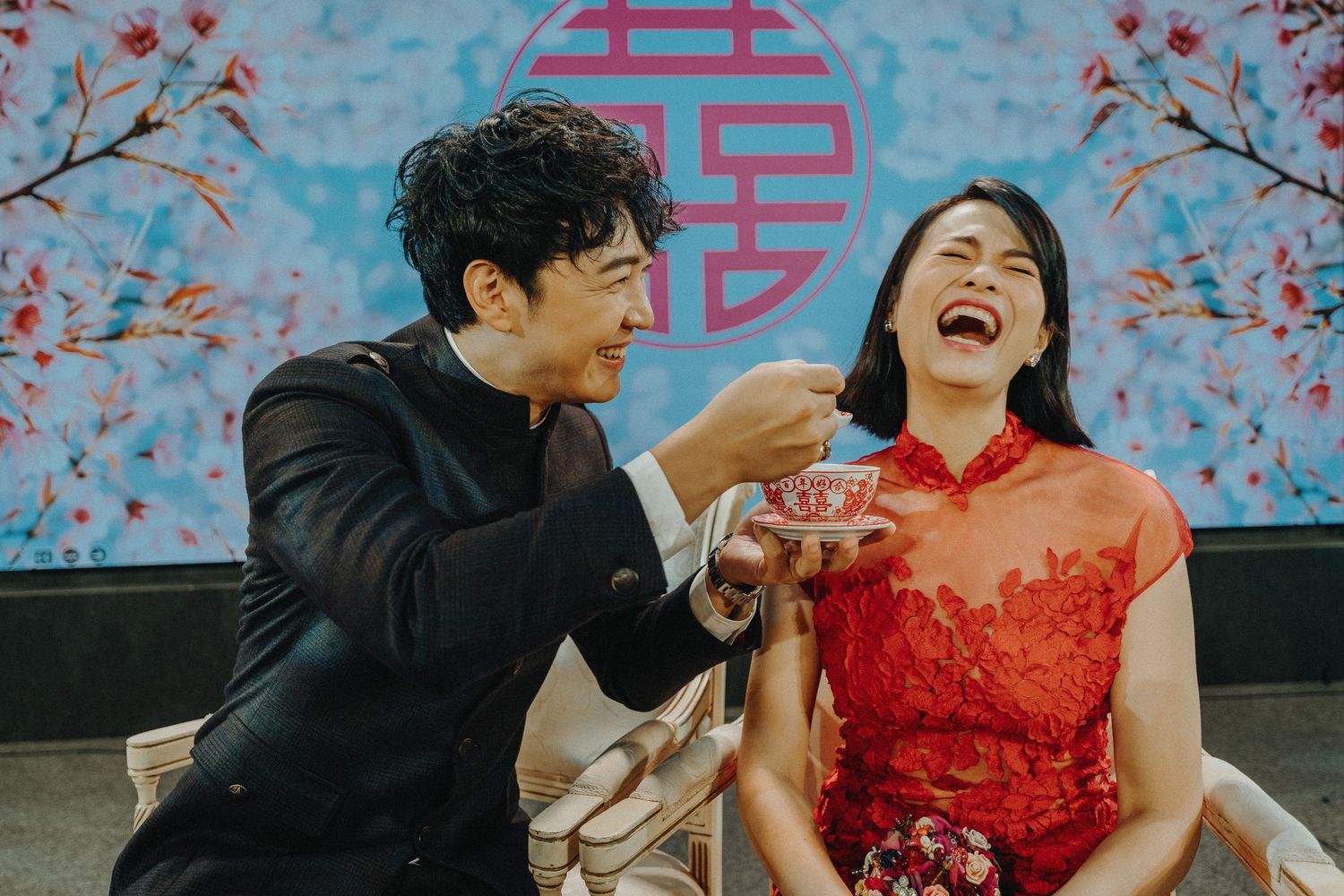 A couple in traditional dress share a joyful moment against a backdrop with cherry blossoms and Chinese symbols.