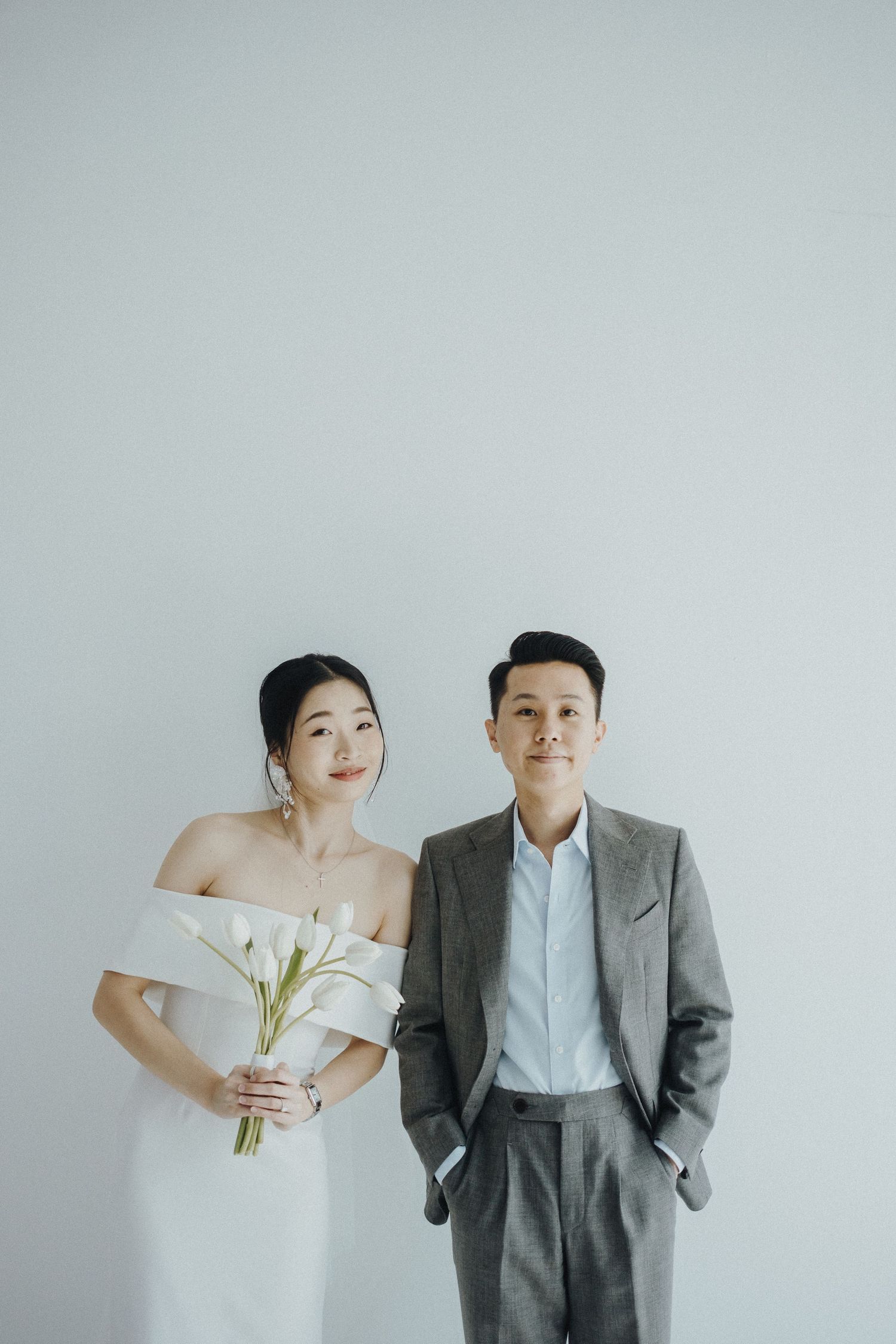 A minimalist wedding photo featuring a bride in white holding calla lilies and a groom in grey suit against light background.