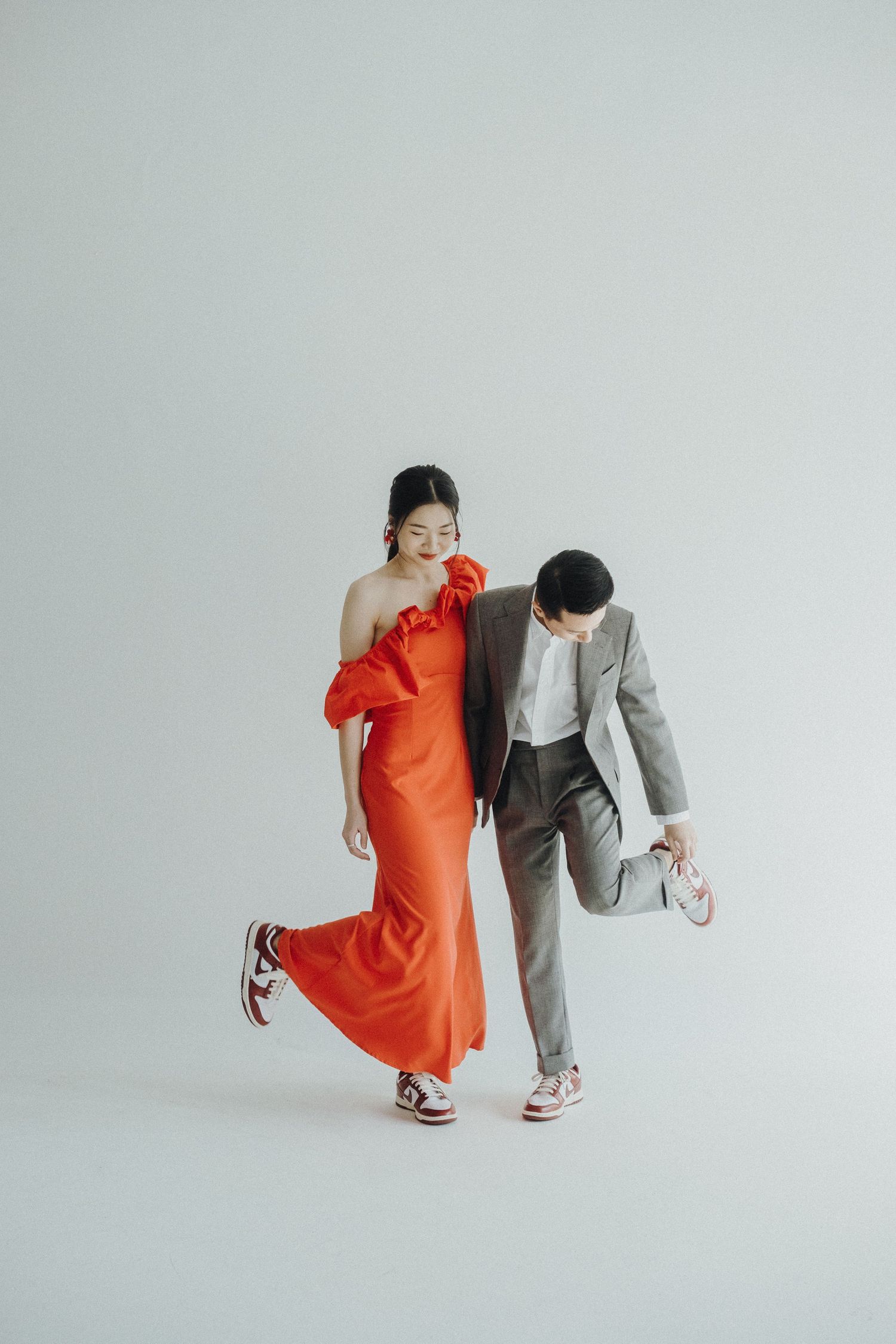 Playful couple in orange dress and gray suit perform dynamic dance moves against white backdrop.