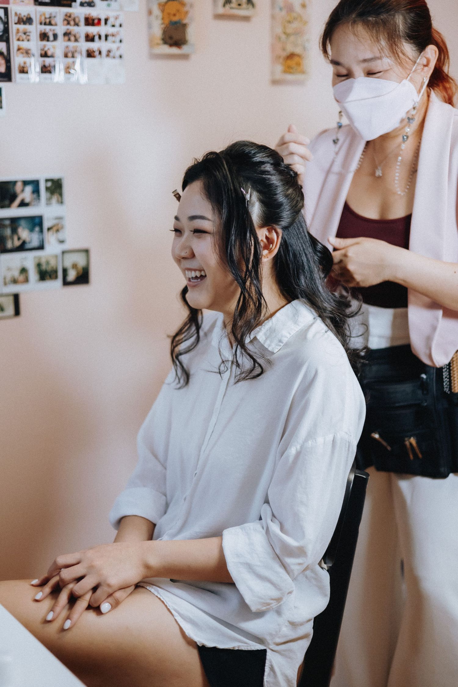 A smiling Asian bride gets her hair styled by a masked hairstylist in a bright salon with photos on the wall.