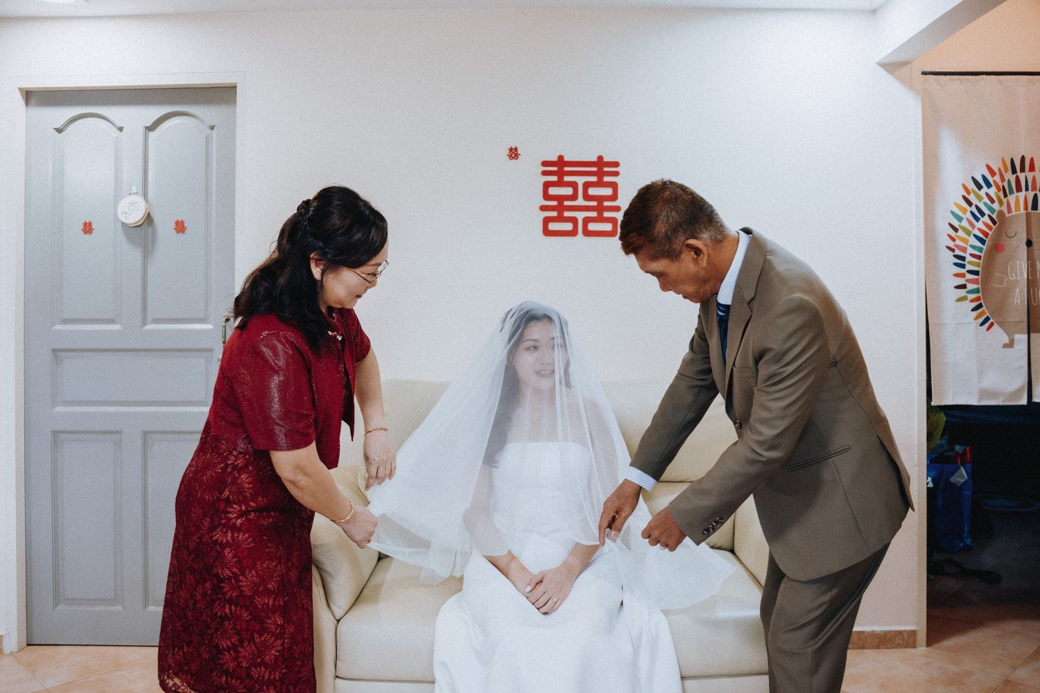 Parents helping a bride in a white wedding dress and veil prepare, with a red Chinese double happiness symbol on the wall.