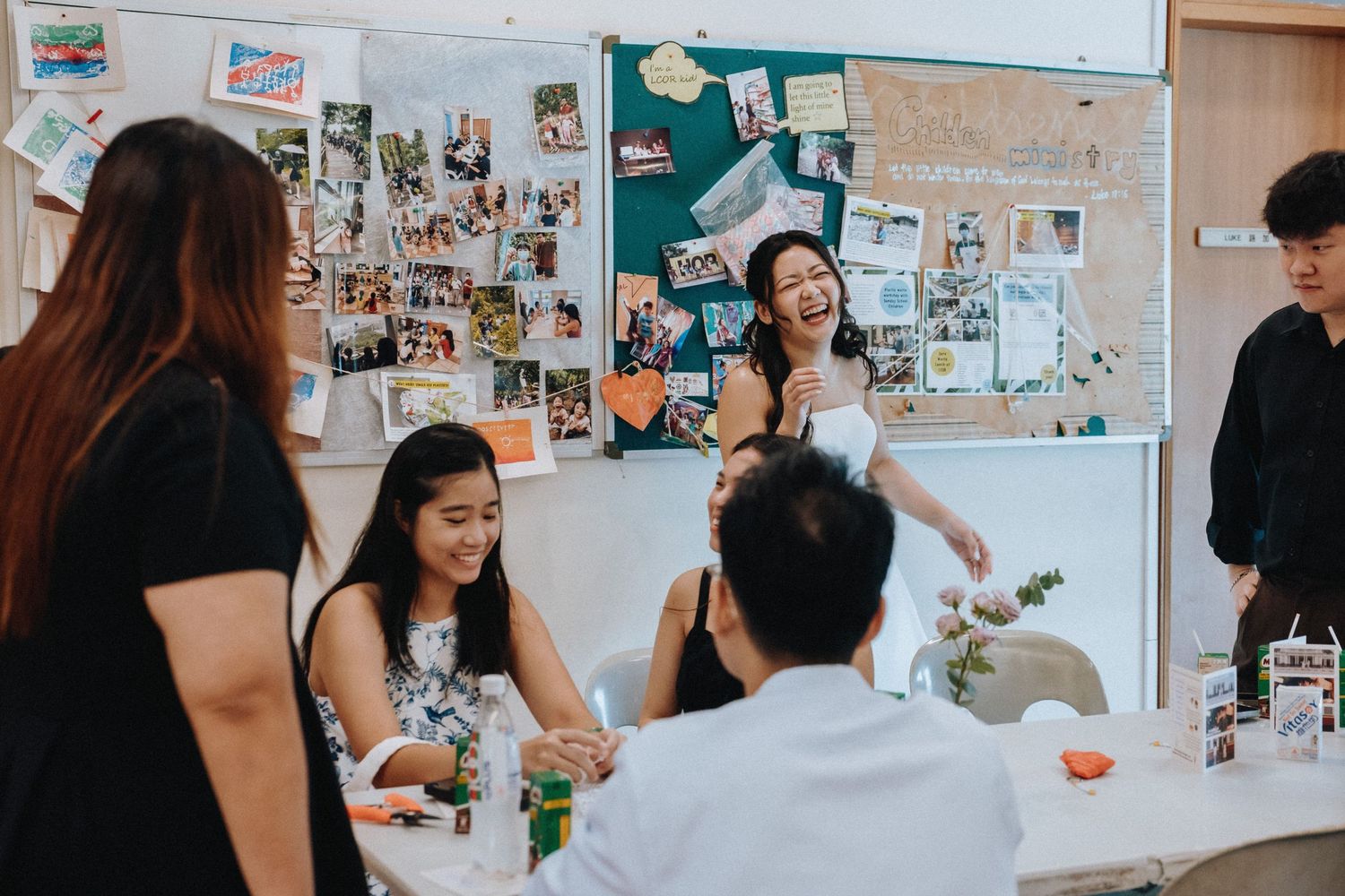 Joyful bride in white dress laughs with guests near a photo memory board at reception.