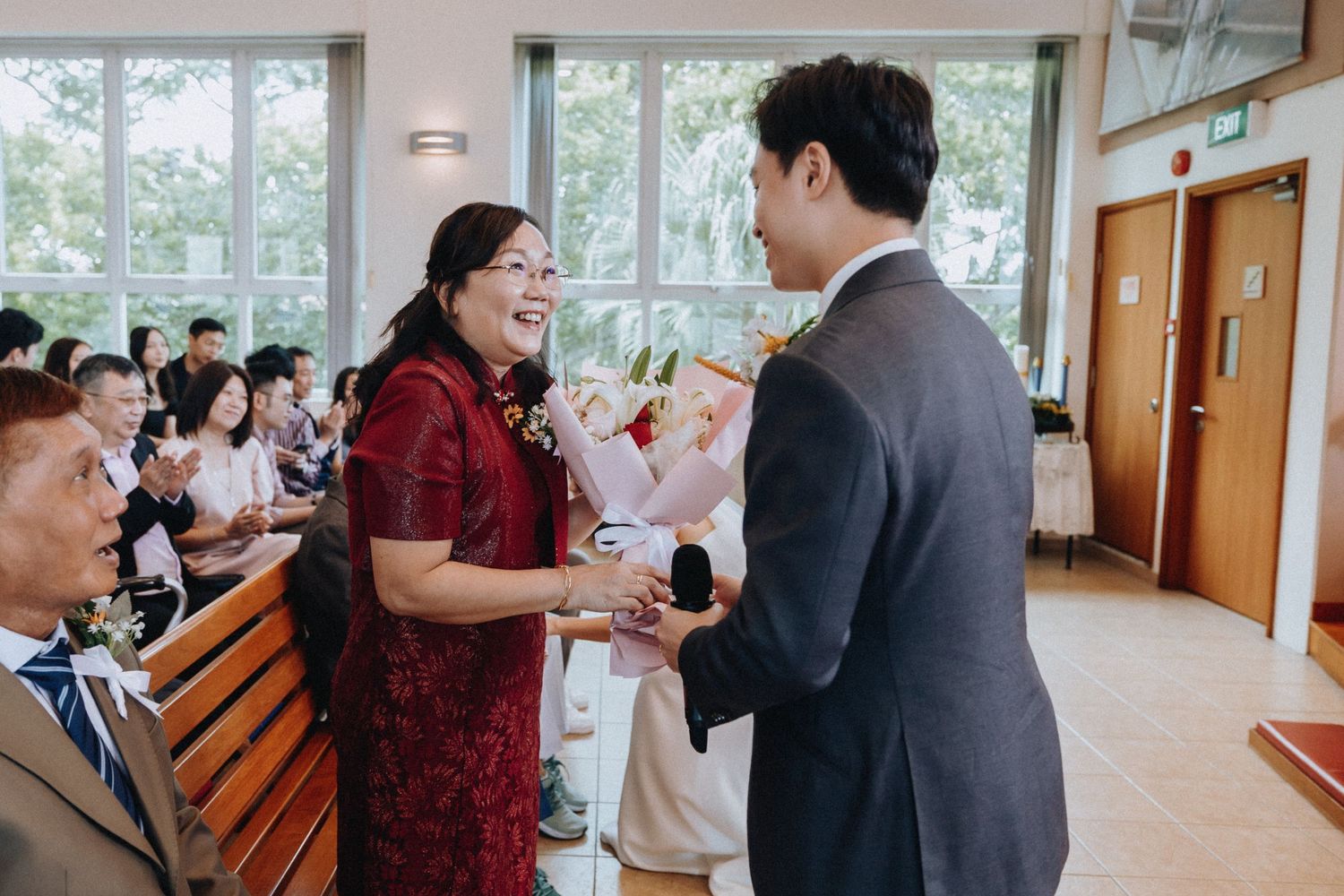 Groom in grey suit presents flowers to a smiling woman in red qipao during a wedding reception.