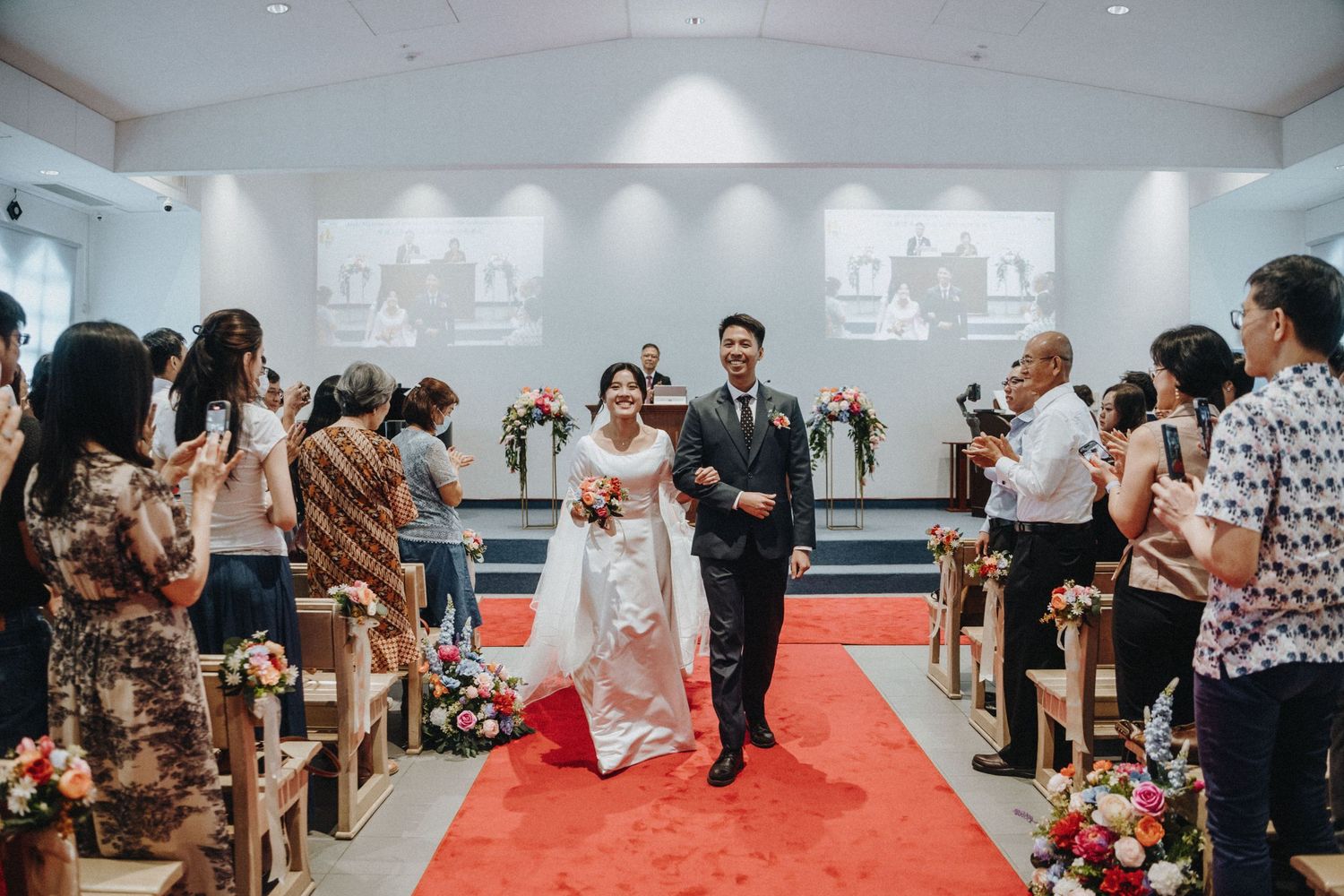 A newlywed couple walks joyfully down a red-carpeted church aisle as guests applaud.