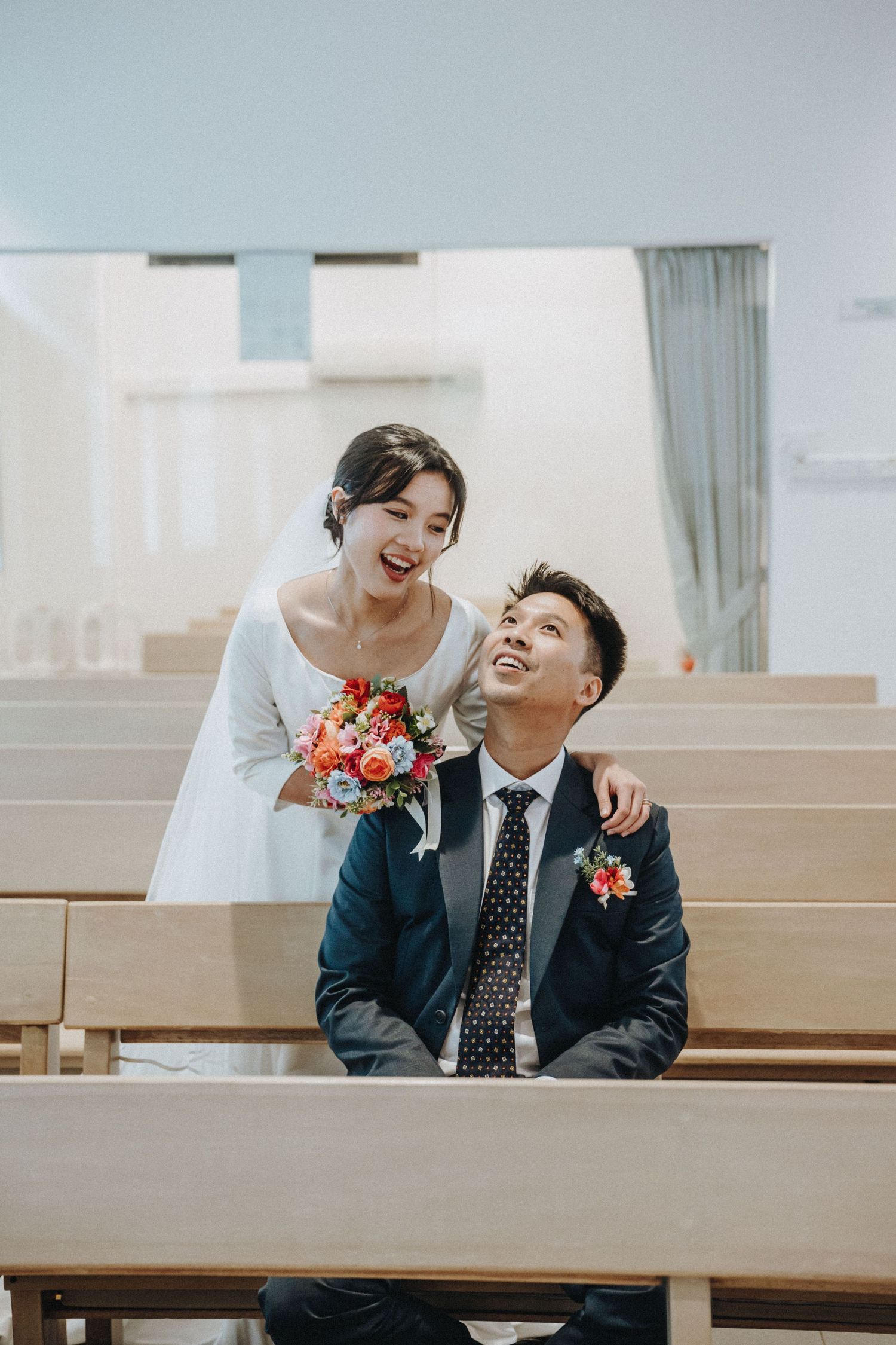 Laughing bride leaning over groom seated on church pew holding colorful wedding bouquet.