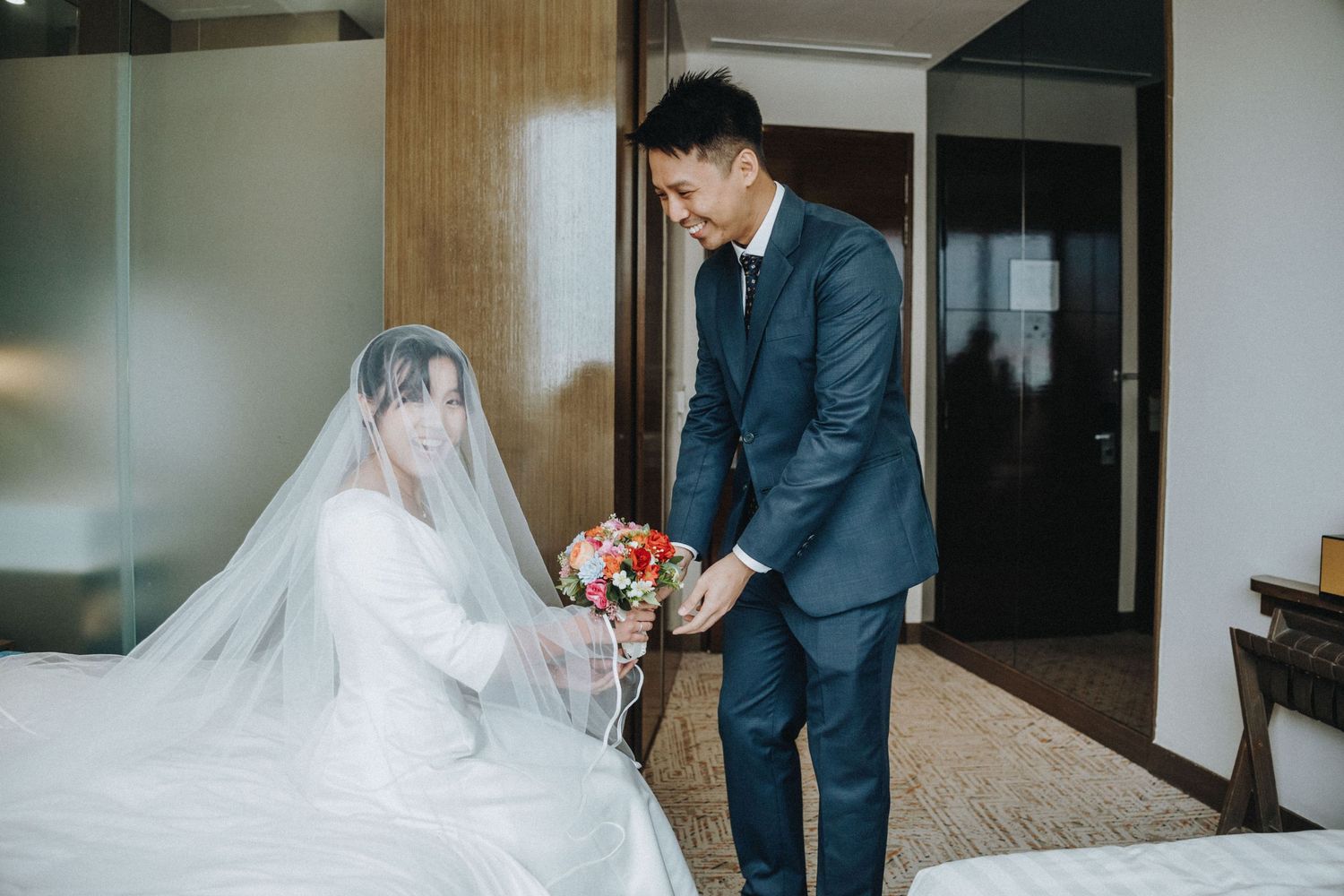 Groom in navy suit presenting colorful bouquet to bride in white gown and veil in elegant hotel room.