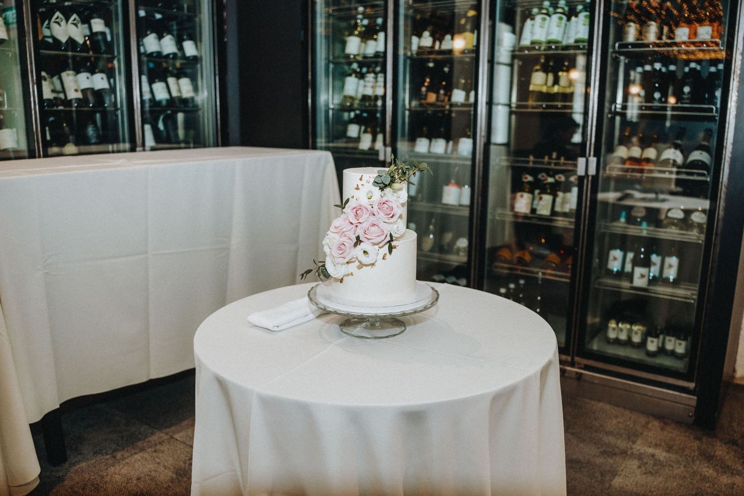 Elegant two-tier white wedding cake with pink roses on a round table in front of wine display fridges.