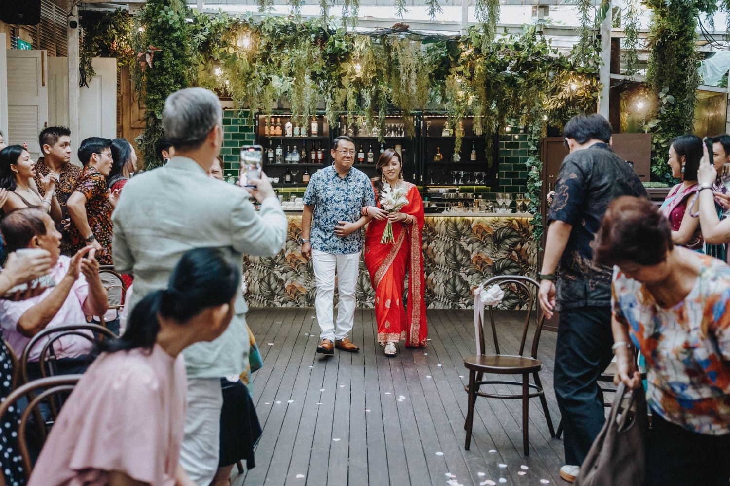 Bride in red saree walks down the aisle with escort at a lush garden wedding ceremony.