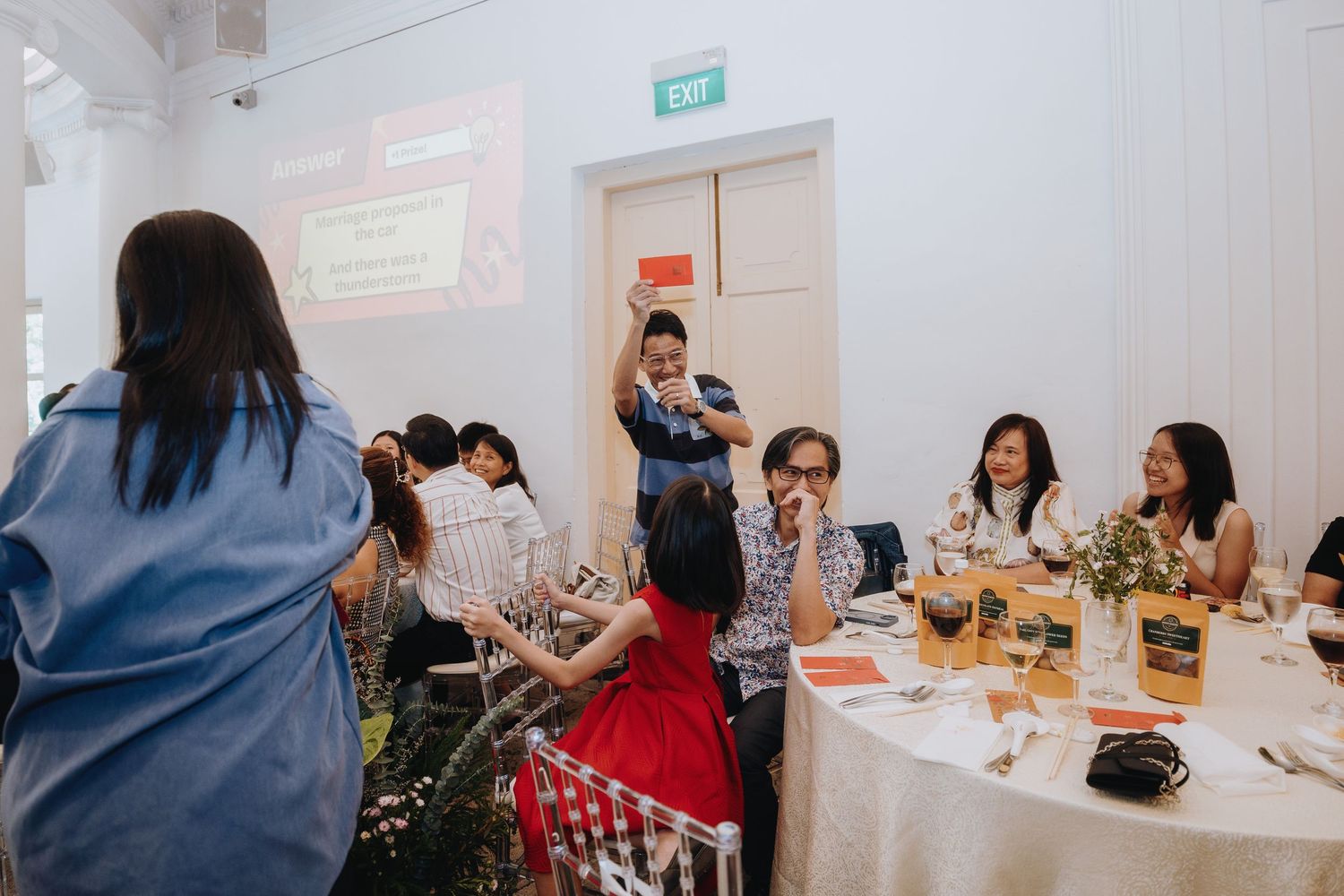 Wedding guests laugh and interact at decorated reception tables while host distributes red envelopes.