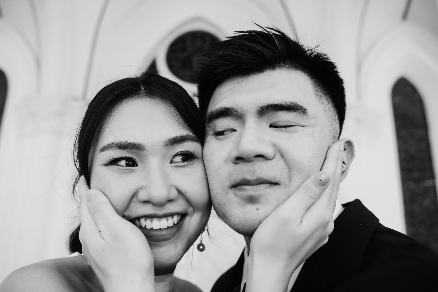 Black and white close-up of a smiling bride and groom posing playfully outside a church.