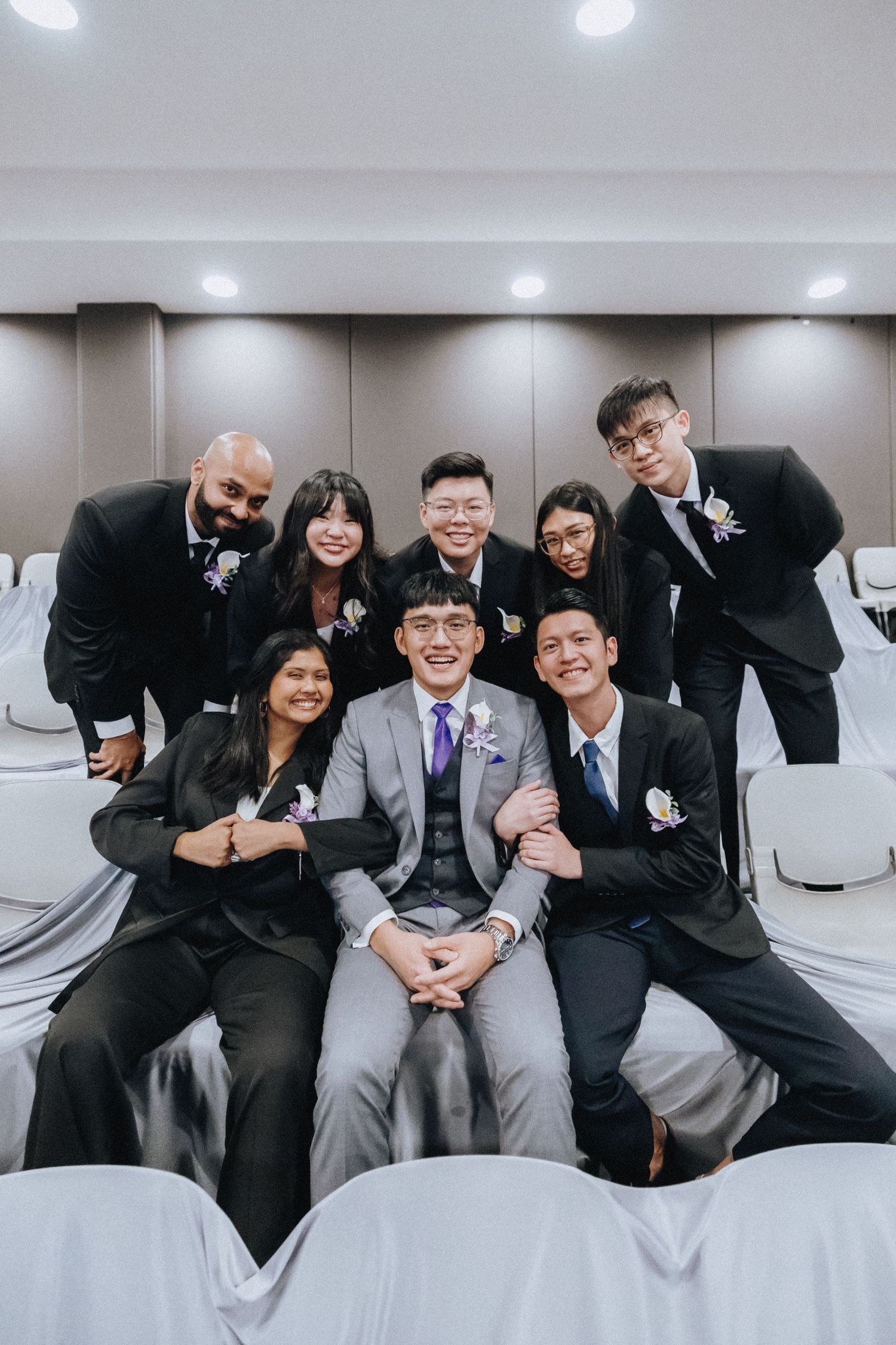 Group photo of wedding party members posing together in formal attire against white chairs.